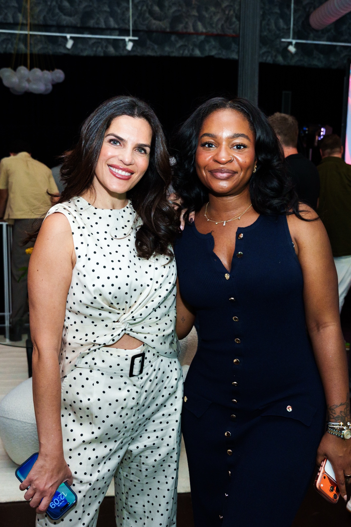 Two women pose together in a dim lounge, one in a polka dot jumpsuit and one in navy