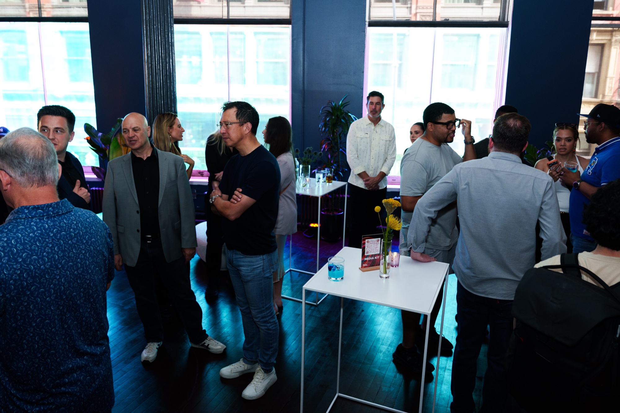 Guests chat around a white cocktail table in a loft with tall windows and a yellow flower in a vase