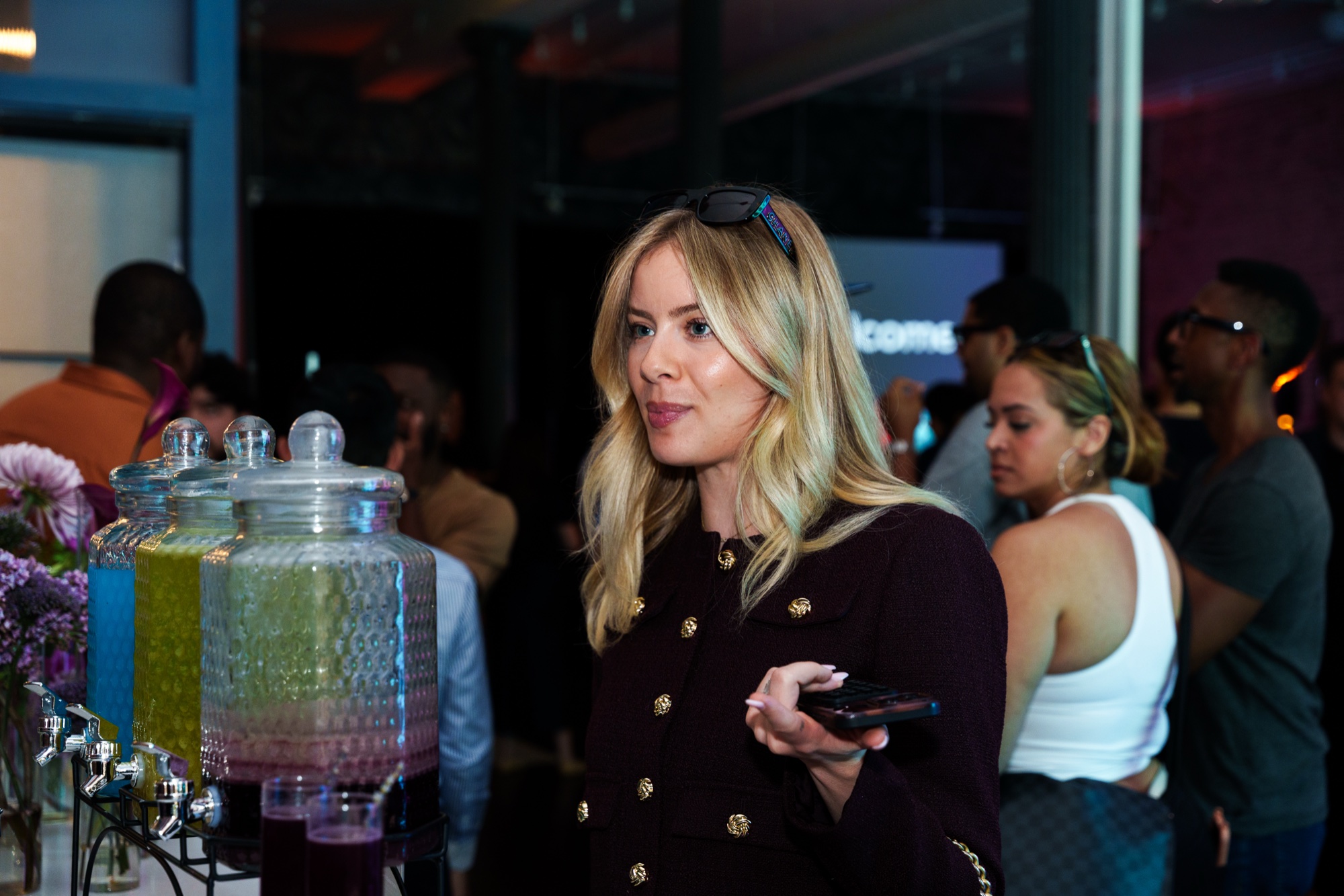 Blonde woman in a black jacket holds a phone beside glass drink dispensers of lime and purple juice