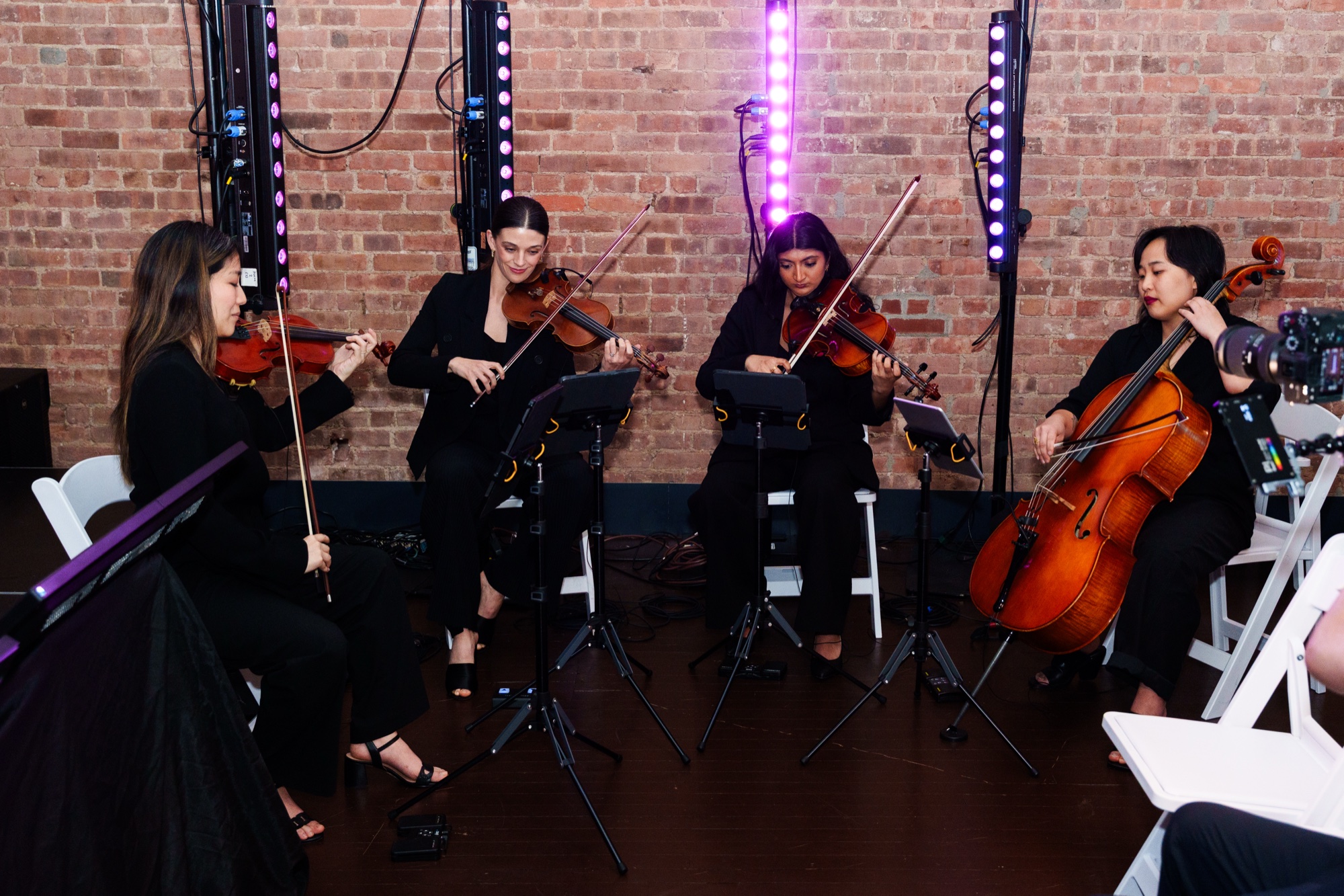 String quartet in black plays violins, viola and cello in front of a brick wall lit by purple lights
