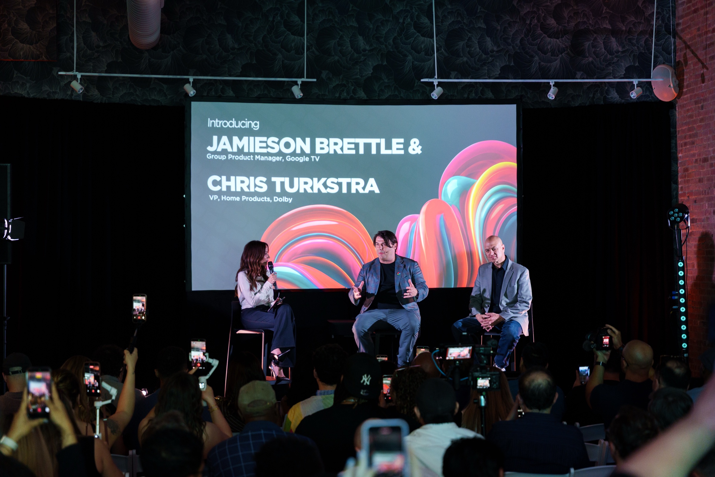 Three speakers on stage under a screen introducing Jamieson Brettle and Chris Turkstra, the audience filming on phones