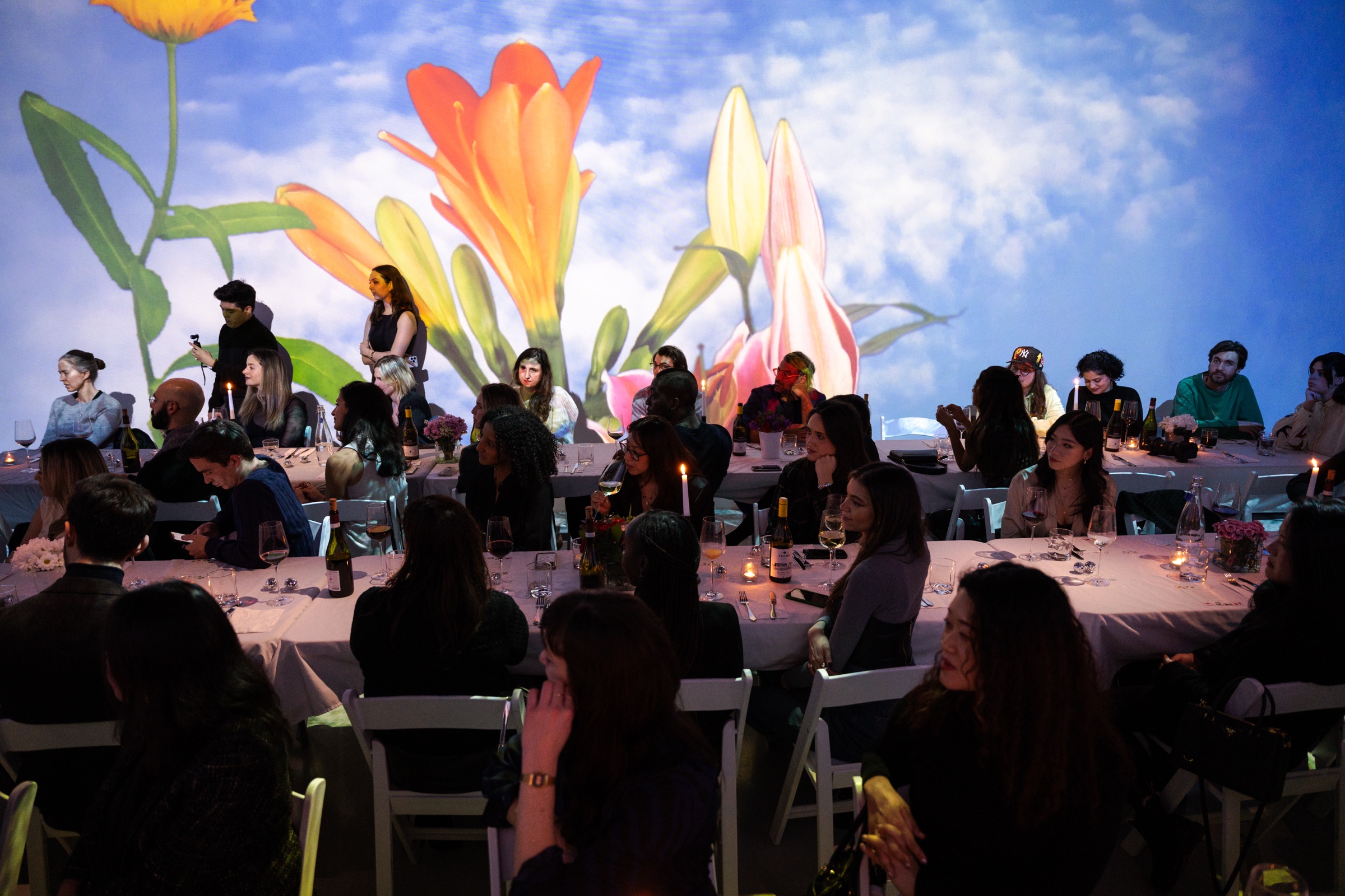 Diners at white tables surrounded by projected blue skies and giant flowers at The Dinner Table