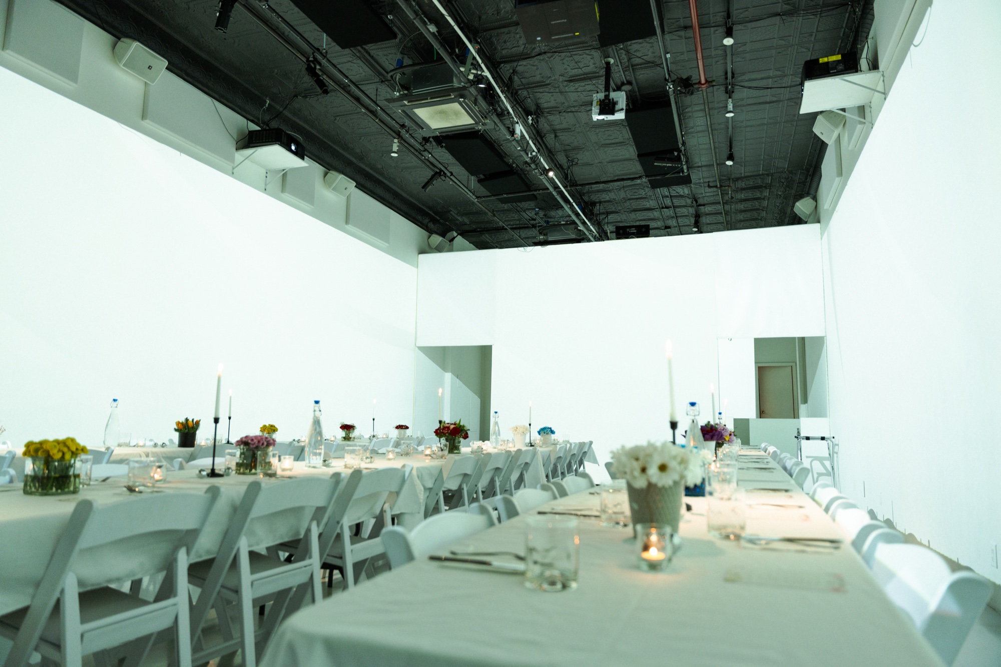 Long white tables with folding chairs, flowers and candles in a bright white walled studio