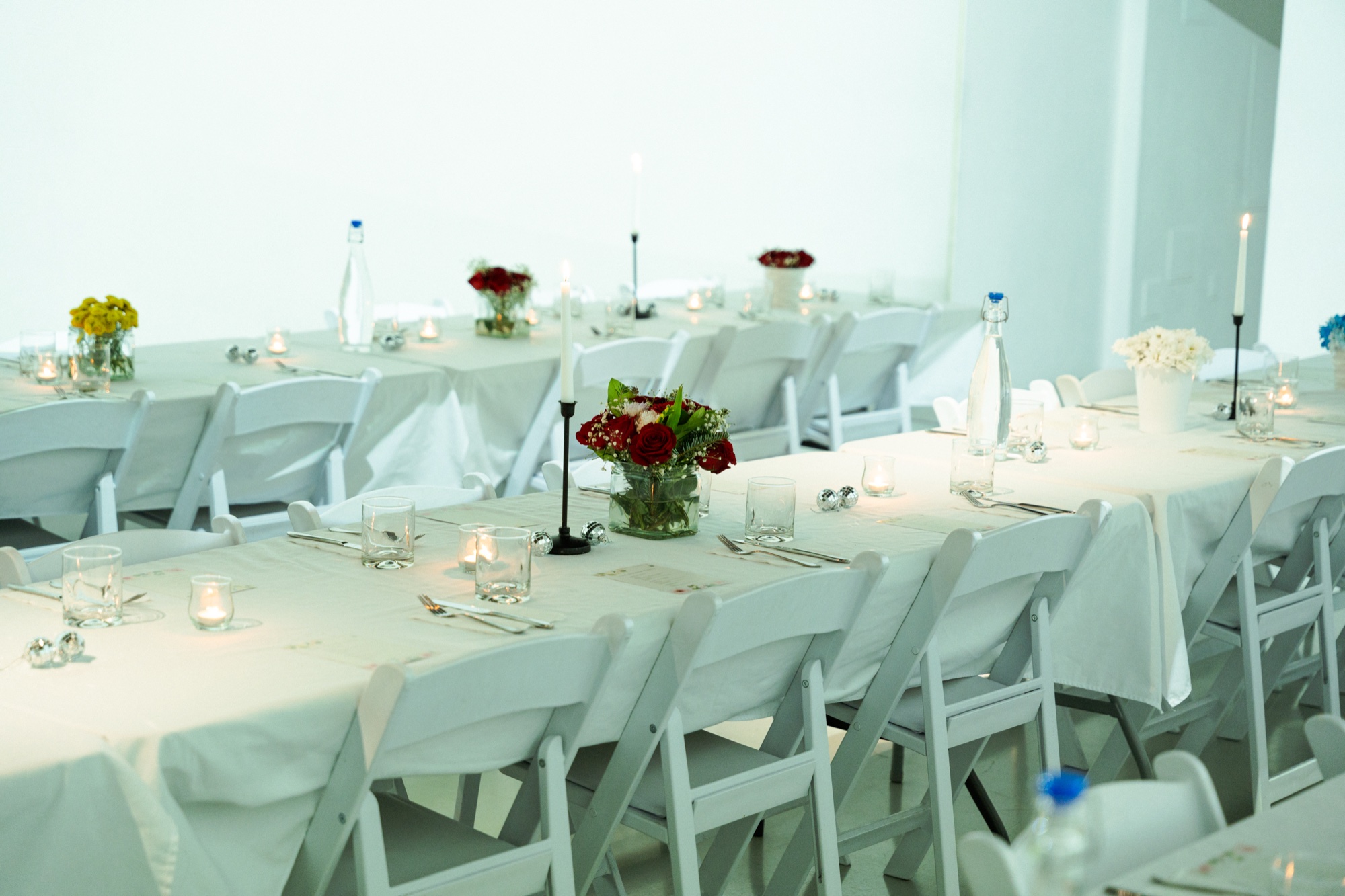 Long white tables with white folding chairs, red flowers, candles and glasses in a white room