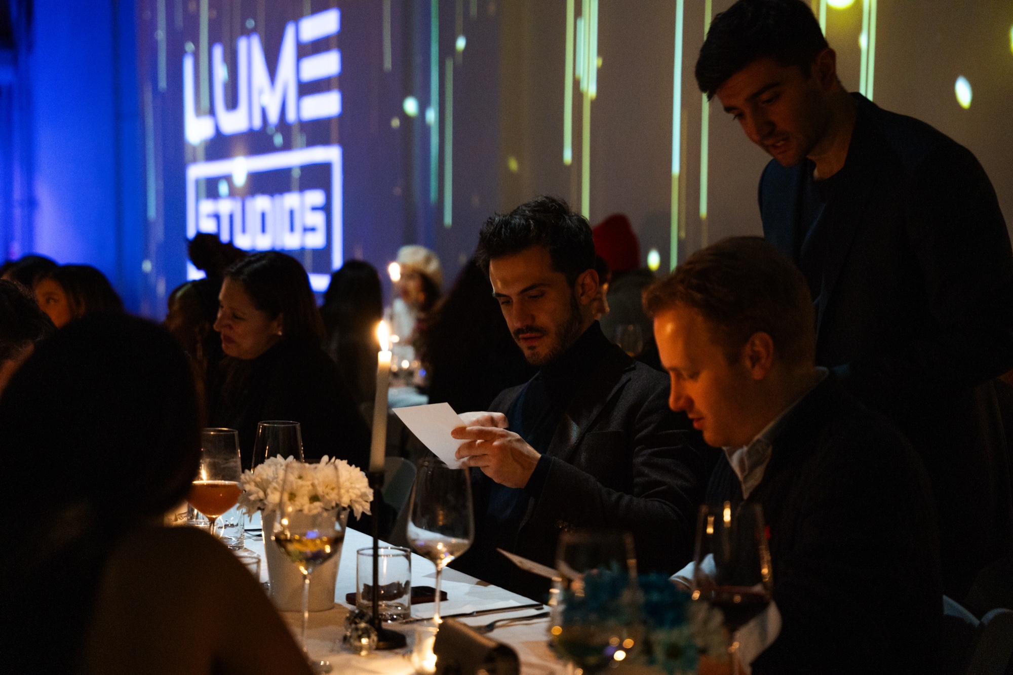 Guests read menus at a candlelit dinner table beneath a wall projection of the Lume Studios logo