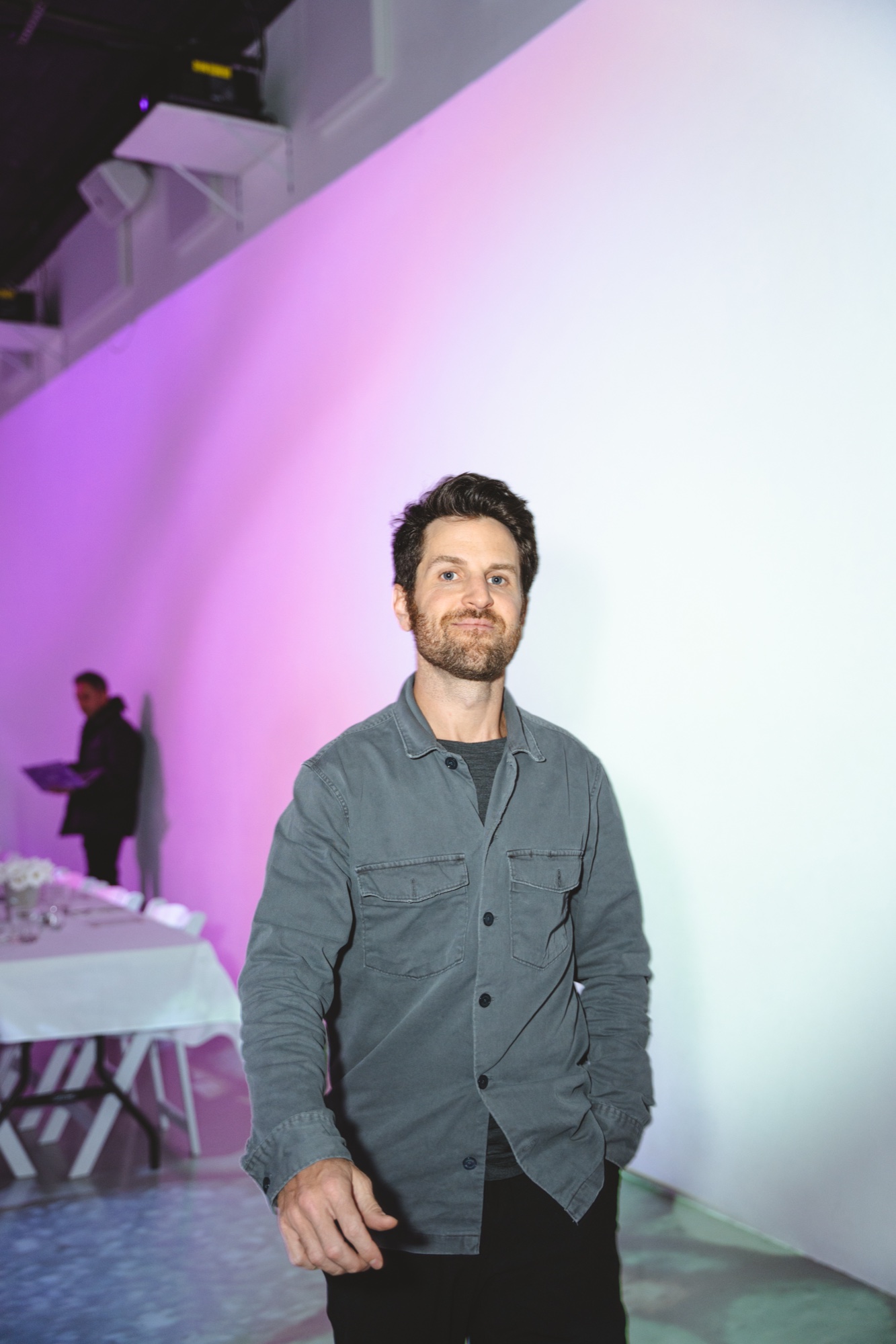 A bearded man in a grey shirt jacket smiles in a white room washed with purple light