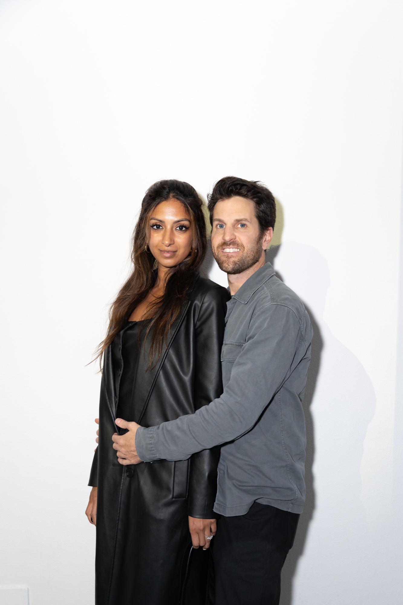 Couple in a black leather coat and a grey shirt posing together against a plain white wall