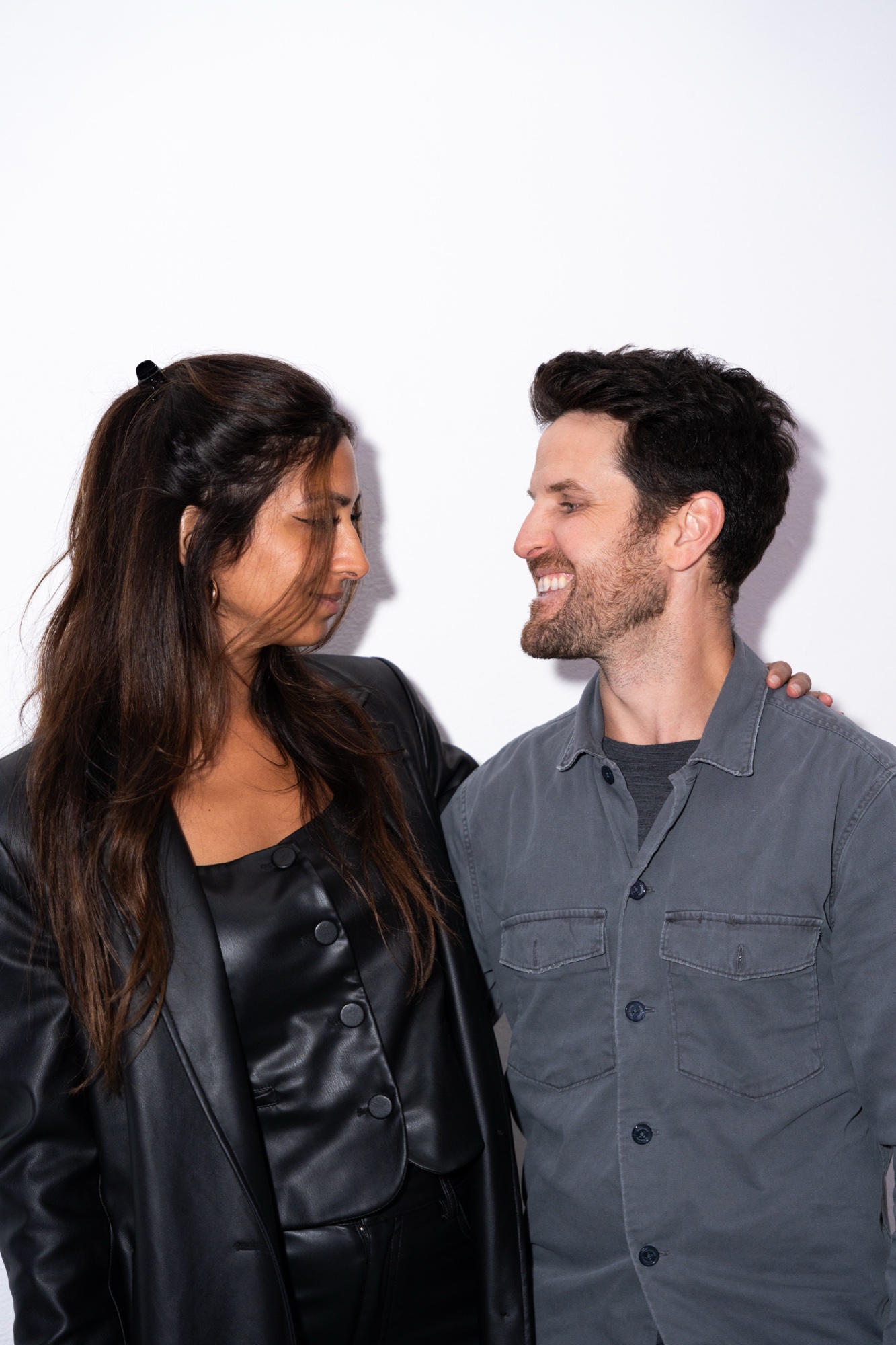 A woman in a black leather jacket and a smiling man in a grey shirt pose against a white wall