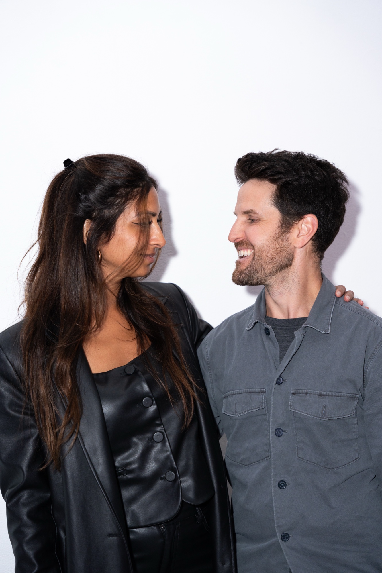 A woman in a black leather jacket and a smiling man in a grey shirt stand against a white wall