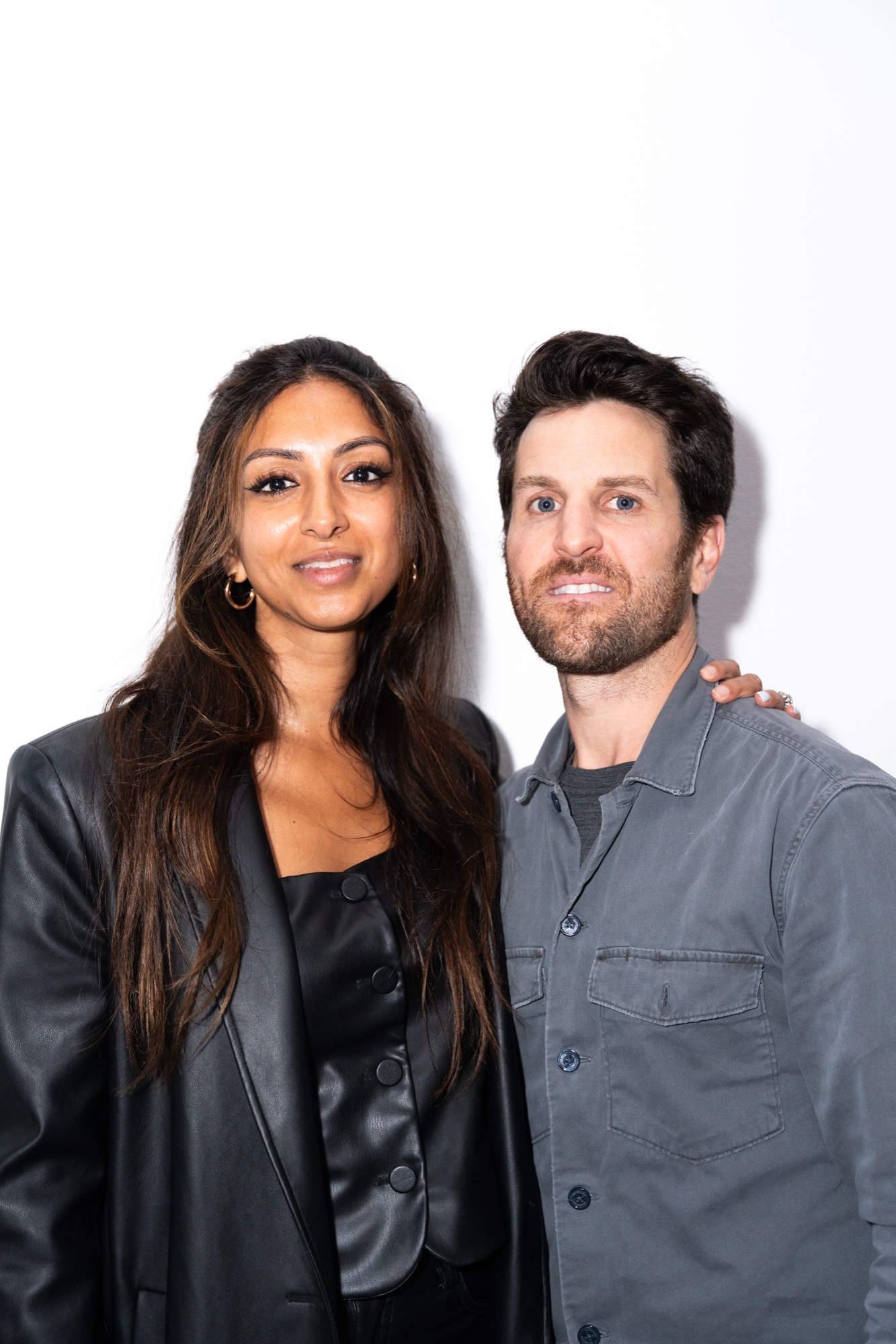 A woman in a black leather blazer and a man in a grey shirt jacket pose against a white wall