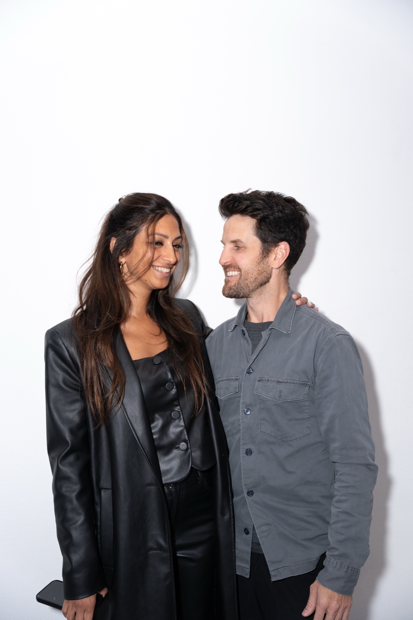 Smiling couple in a black leather coat and grey overshirt sharing a look against a white wall