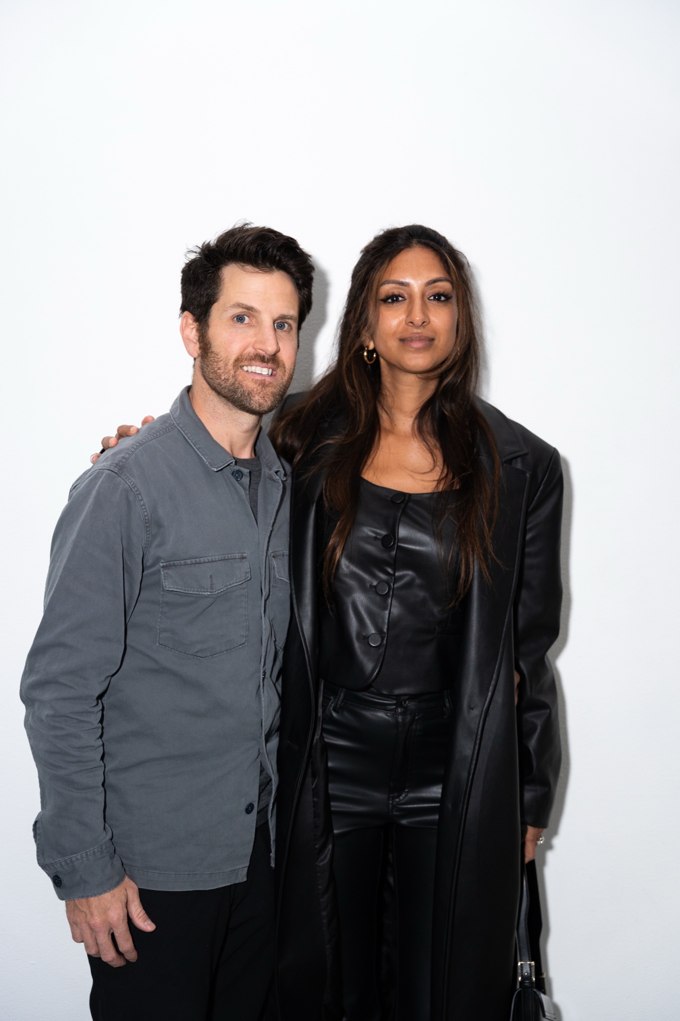 A man in a grey shirt and a woman in a long black leather coat pose against a white wall