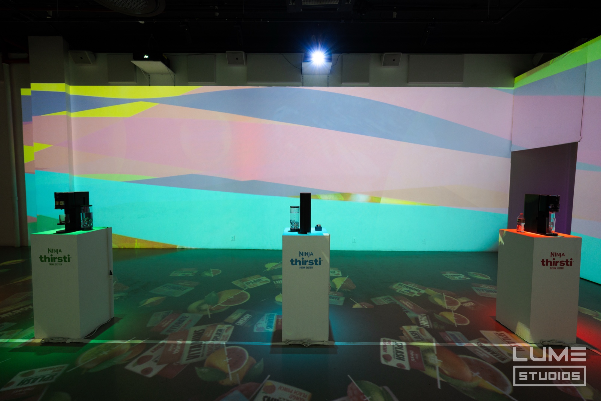 Three white plinths holding Ninja Thirsti drink machines before a wall projected with pastel colour bands