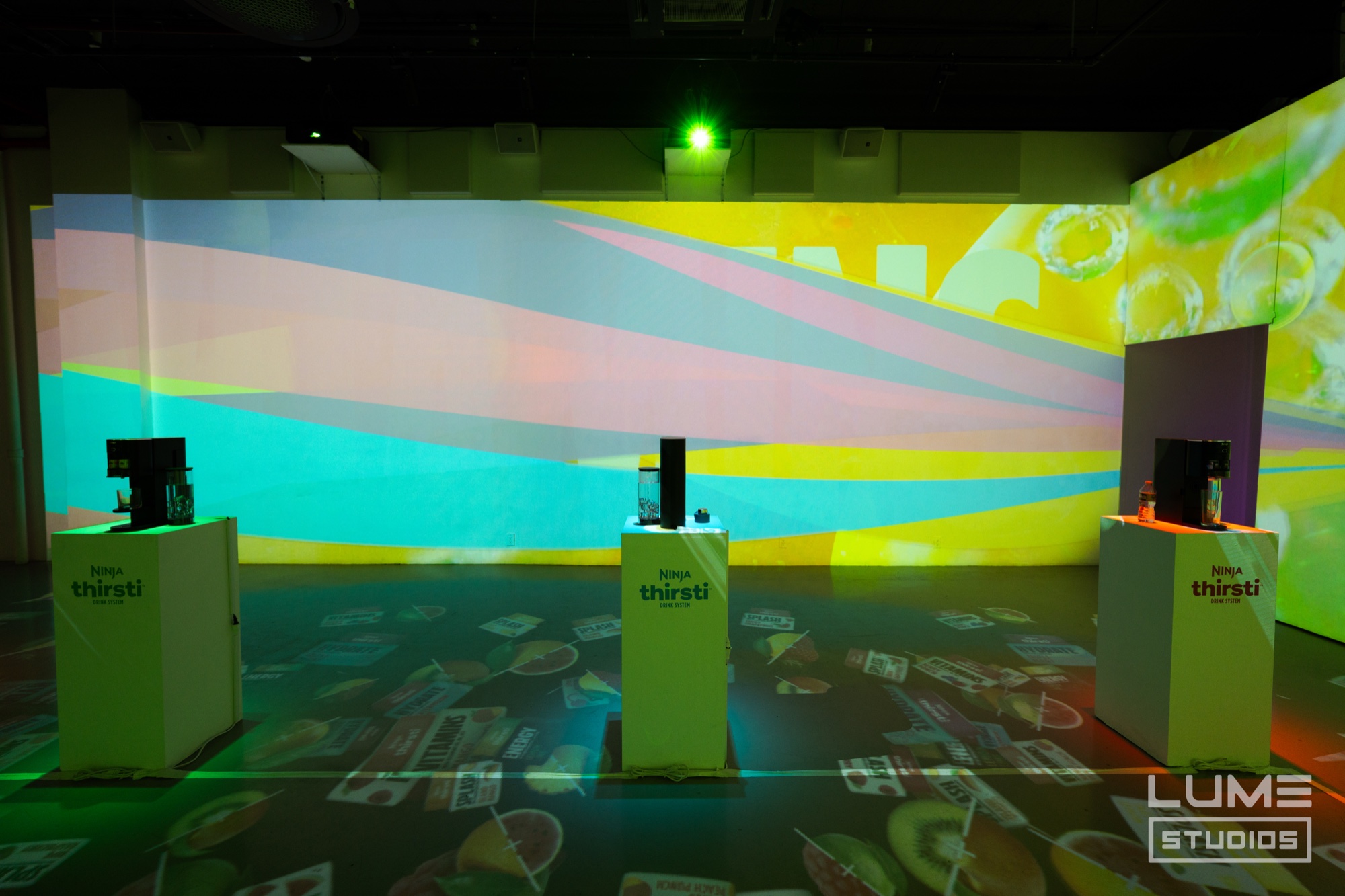 Three white Thirsti plinths before a wall of yellow, teal and pink projected stripes and fruit floor projections