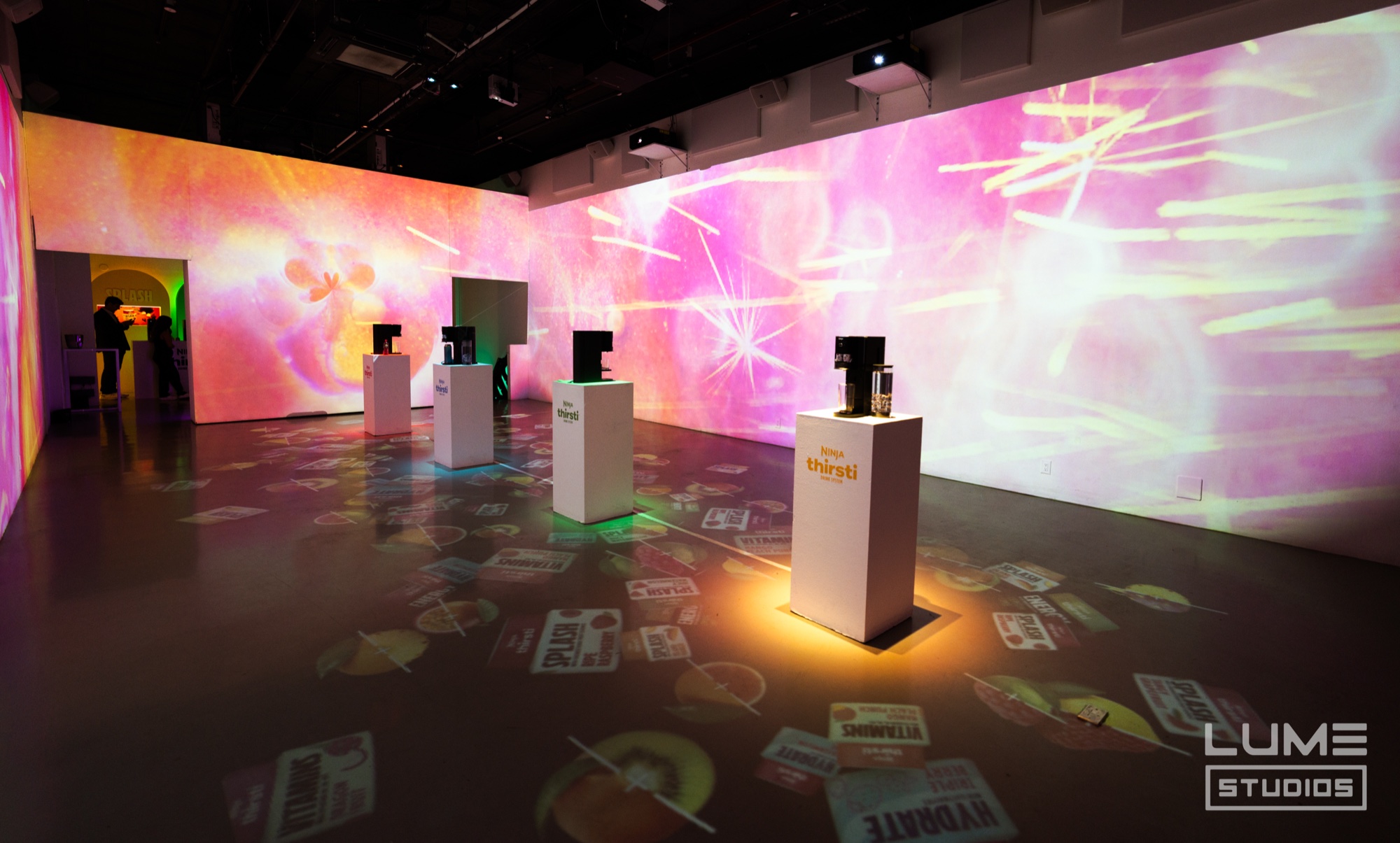 White plinths with drink machines spaced across a room lit by pink and yellow projections of splashing juice