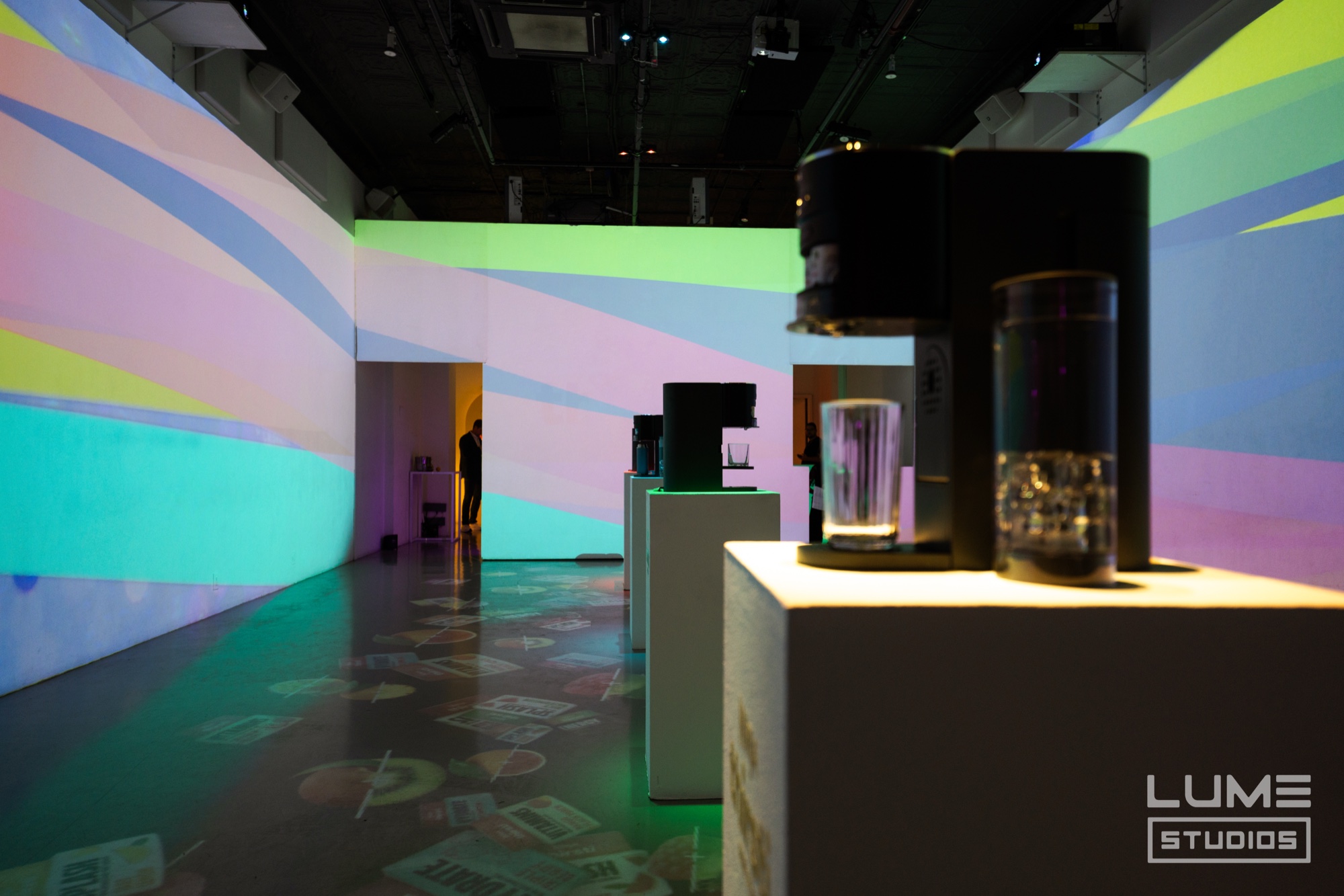 A Ninja Thirsti drink machine and glass on a white plinth in a room projected with pastel stripes