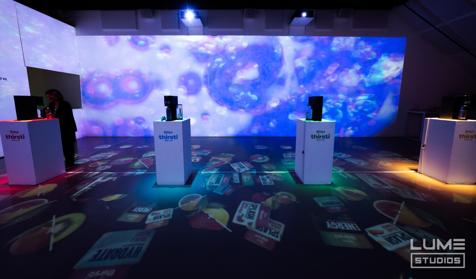 Three Thirsti plinths on a floor of projected drink labels beneath a swirling blue and orange wall