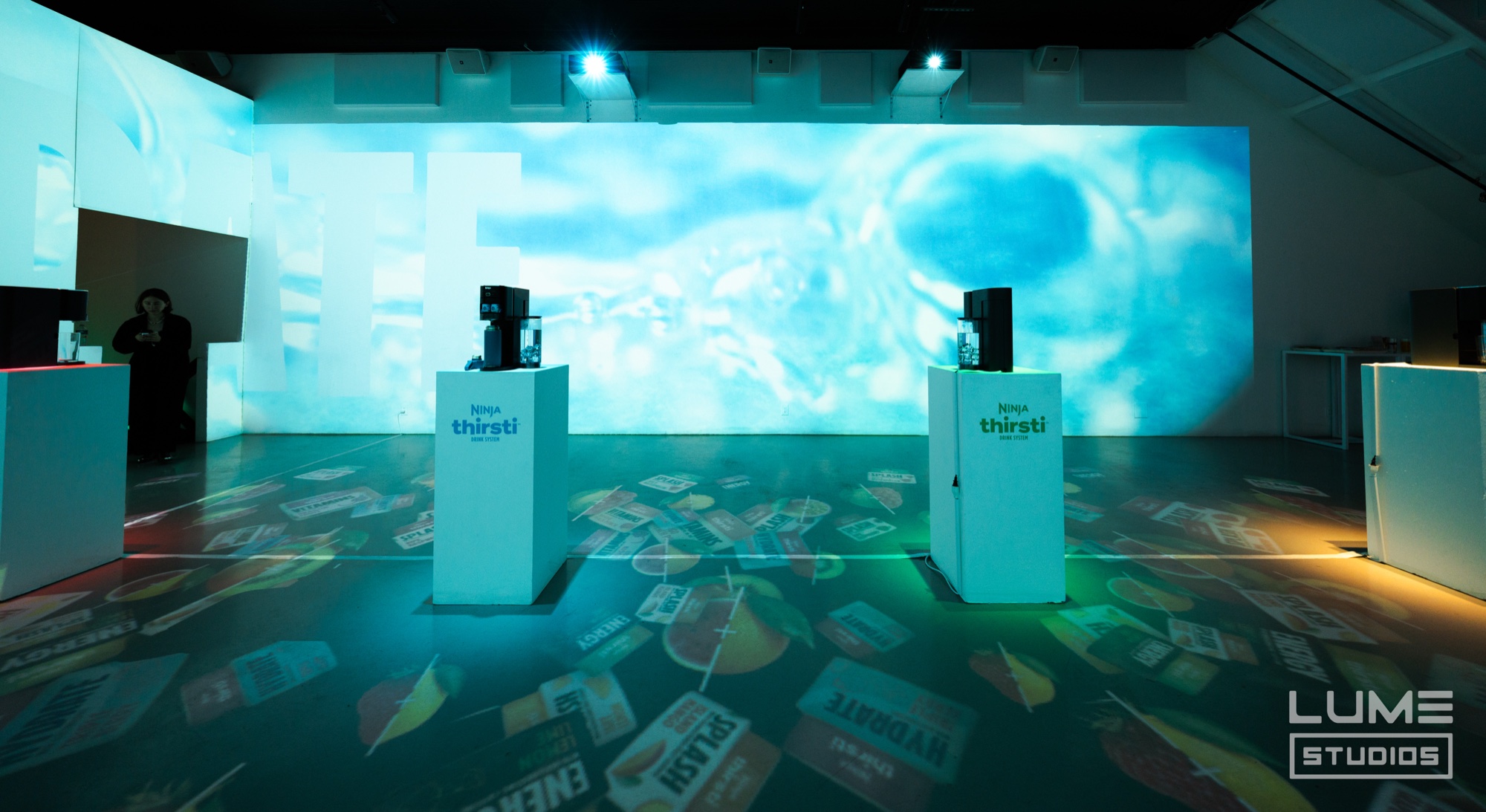 Two white Thirsti dispensers stand before a wall of projected blue bubbles with drink labels scattered across the floor