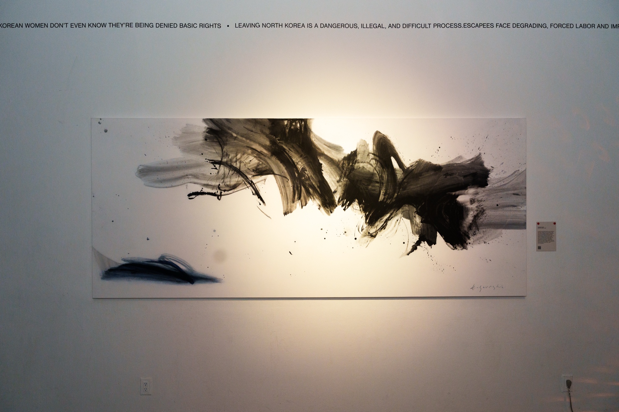 A wide black ink painting of sweeping brushstrokes on a white wall beneath a line of printed text