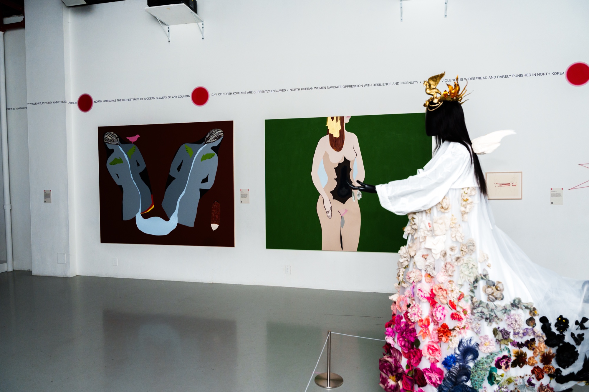A visitor in a white flower covered gown and gold crown views two large figurative paintings in a white gallery