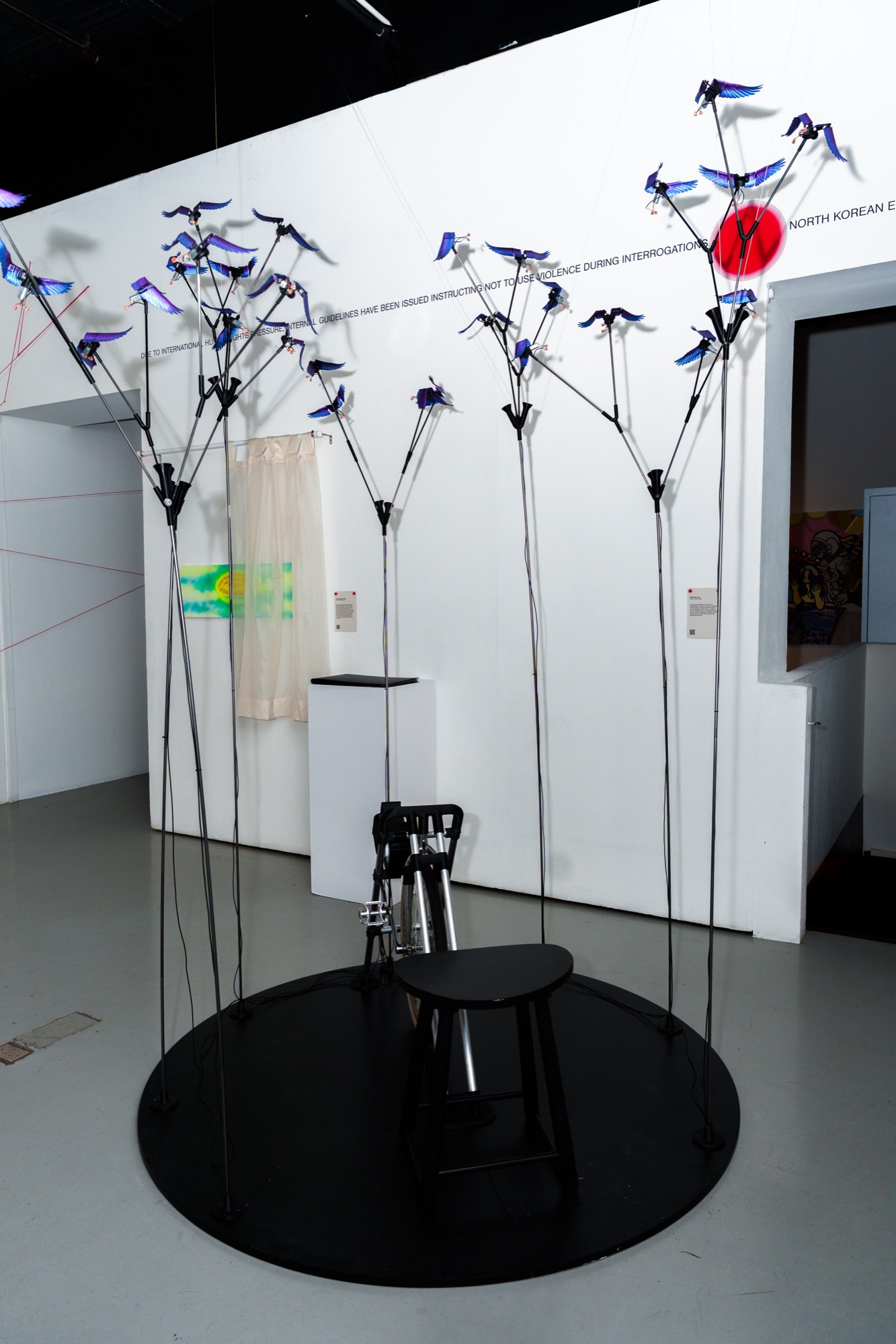 Sculpture of tall black wire stems topped with blue paper birds around a black stool on a round base