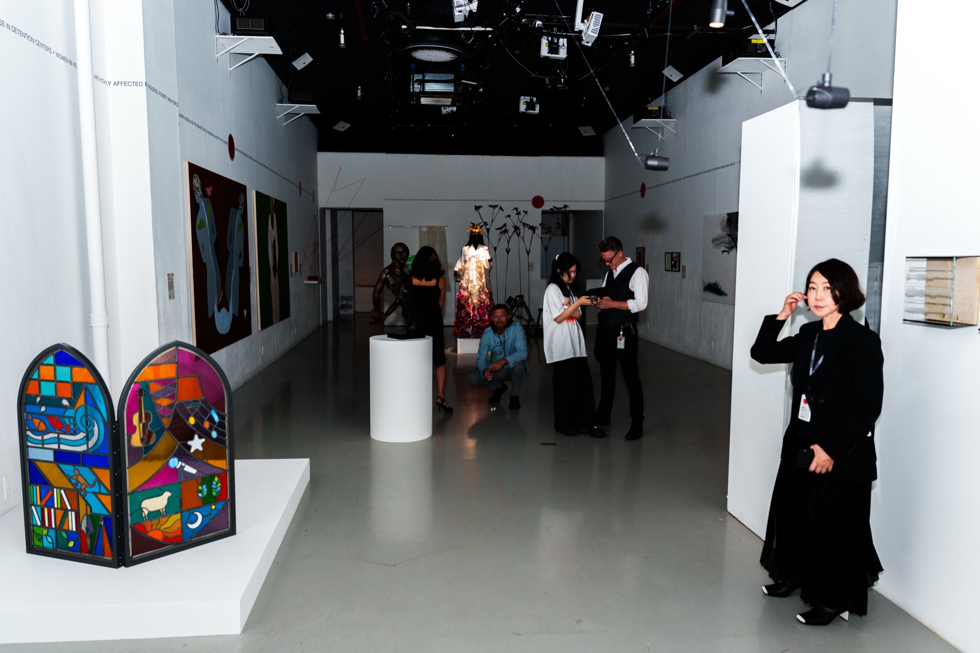 A white gallery with a stained glass style panel in the foreground and guests viewing works at the far end