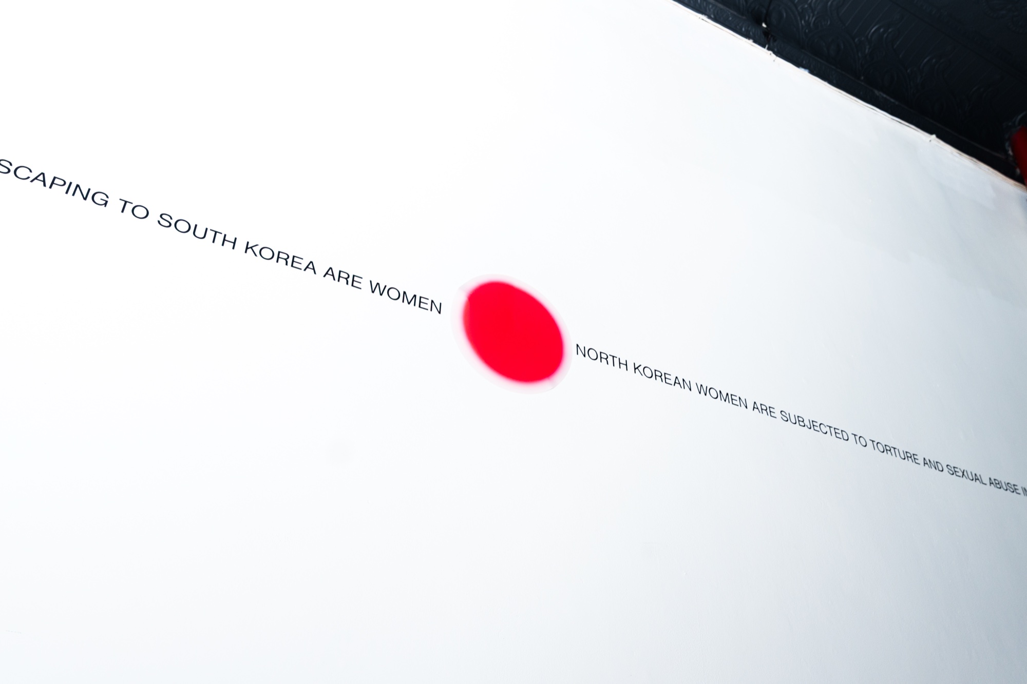 Red dot and thin lines of black text about North Korean women on a white gallery wall