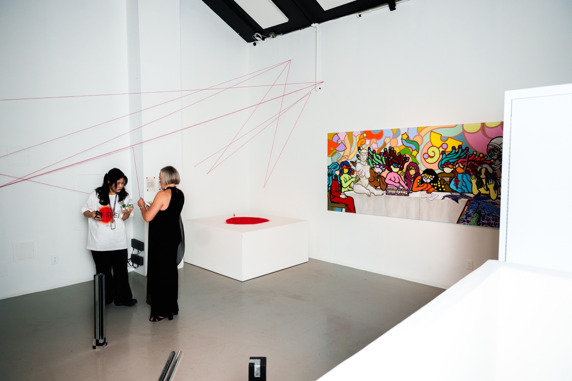 Two women talk in a white gallery under crossing pink strings, a colourful mural on the far wall