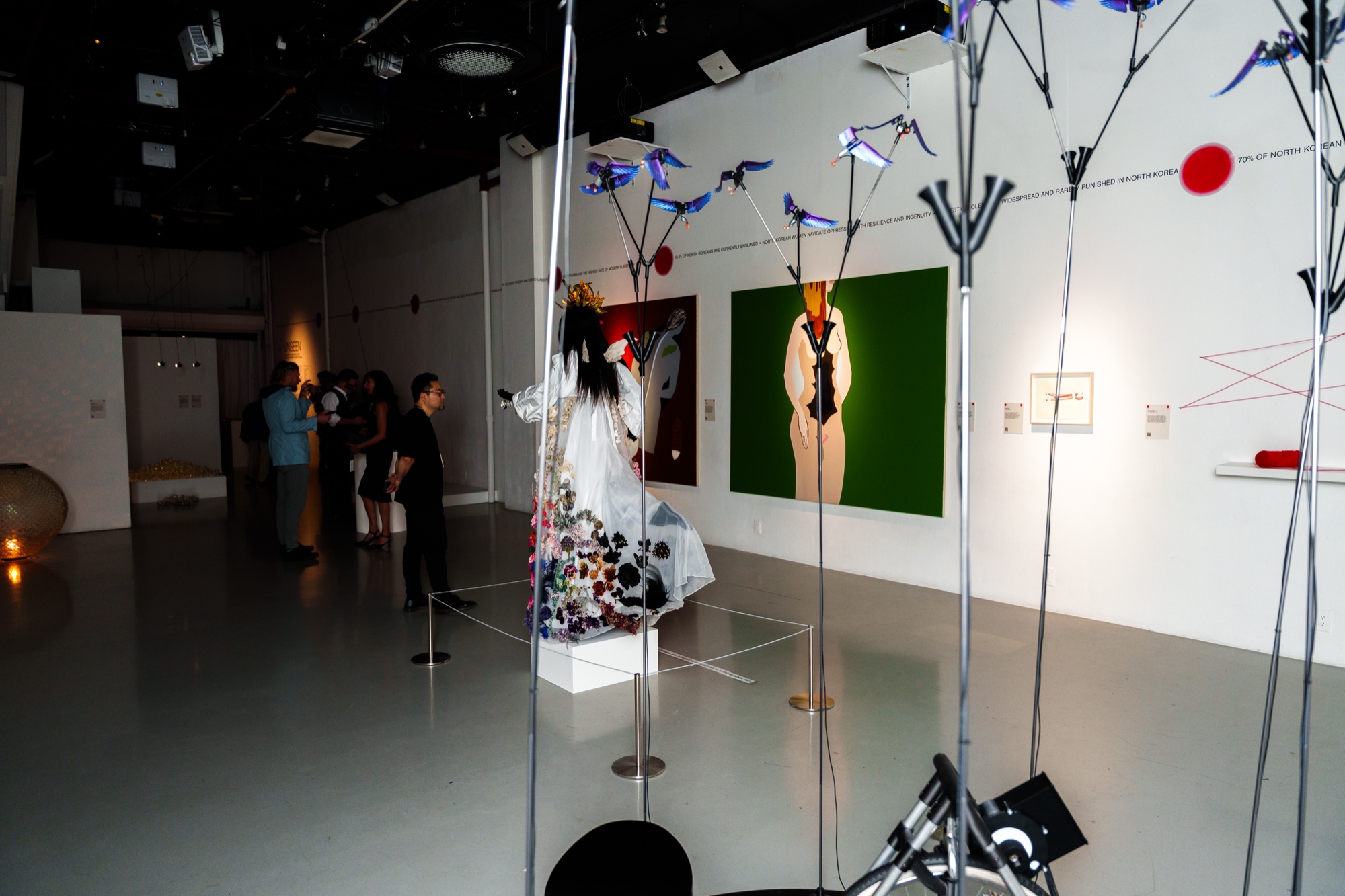 Gallery visitors near a floral gowned mannequin and a green painting, with wire sculptures in the foreground
