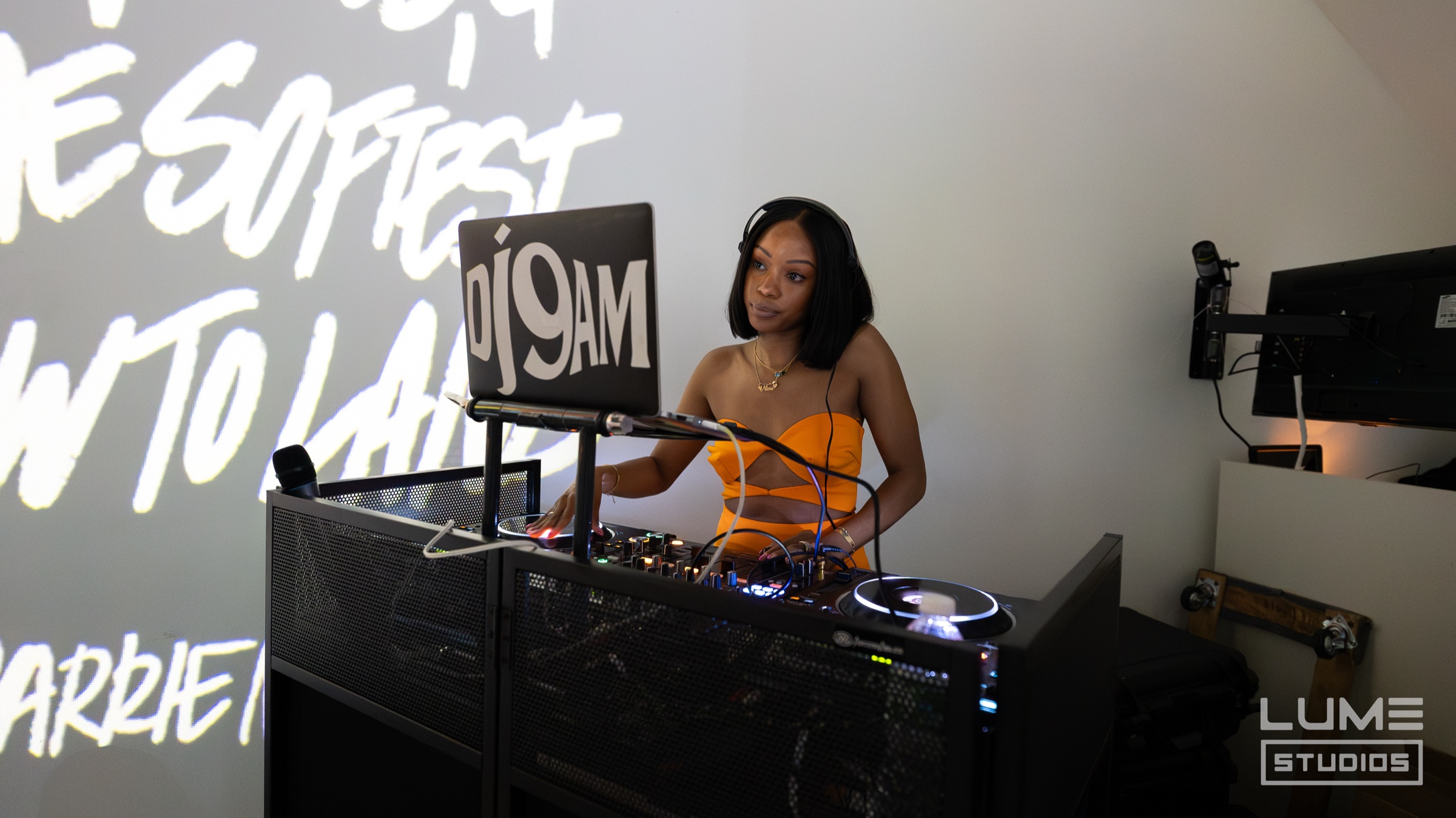 DJ in an orange top plays behind a black booth with a laptop, white script lettering projected behind her