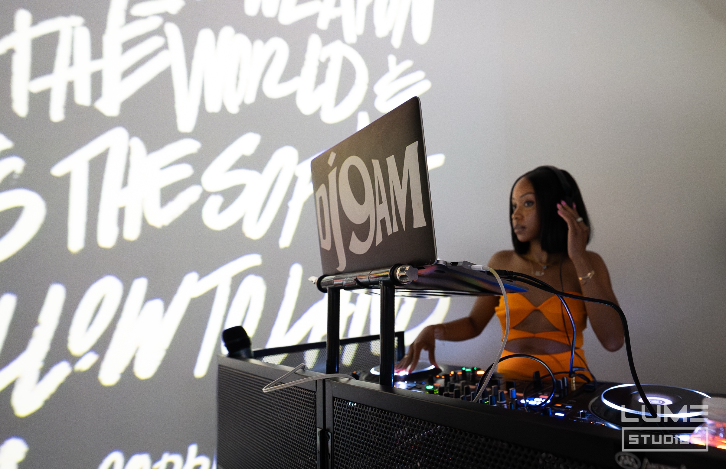 DJ in an orange dress mixes behind a laptop marked DJ 9AM, white graffiti lettering projected behind