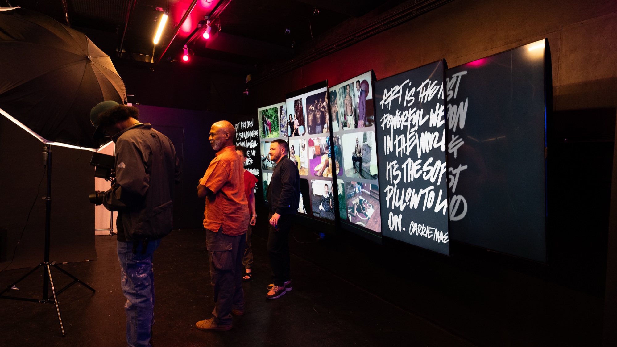 Three guests watch a wall of photo screens and projected white brush lettering under red lights