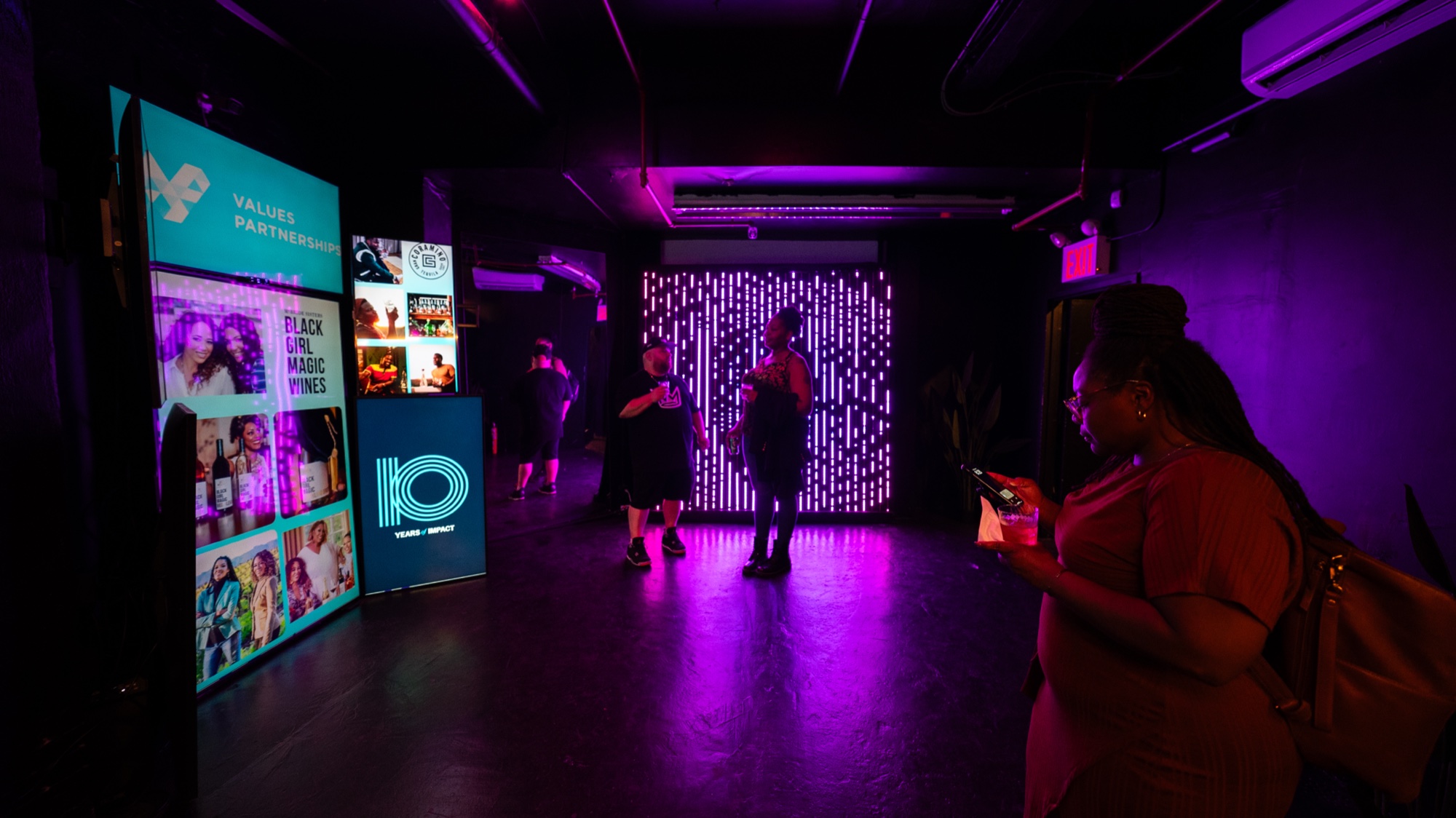 Purple lit hallway with a glowing photo collage panel, a dotted LED wall and guests checking phones