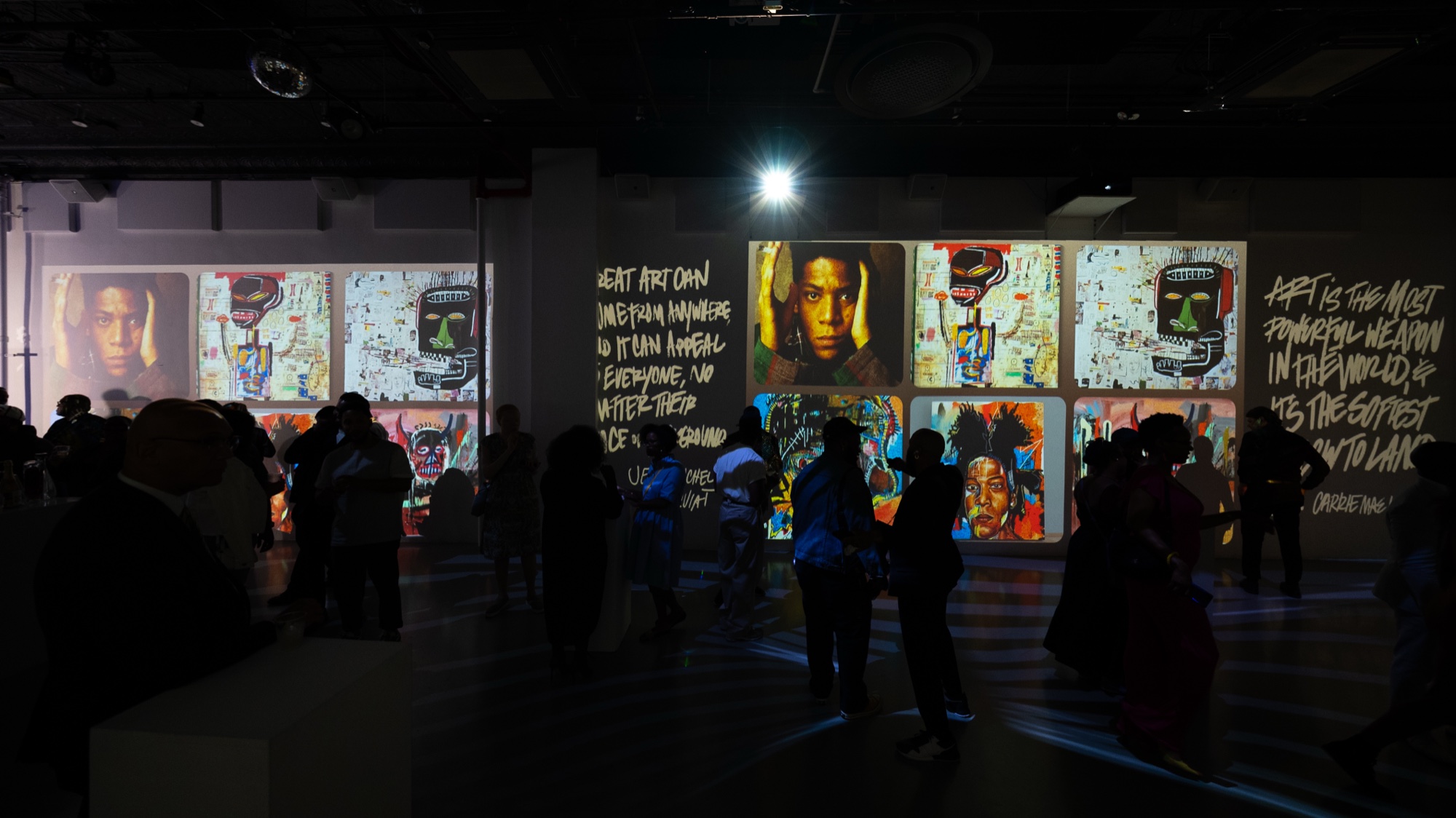 Silhouetted guests view projected colourful portrait paintings and scrawled white quotes on dark walls