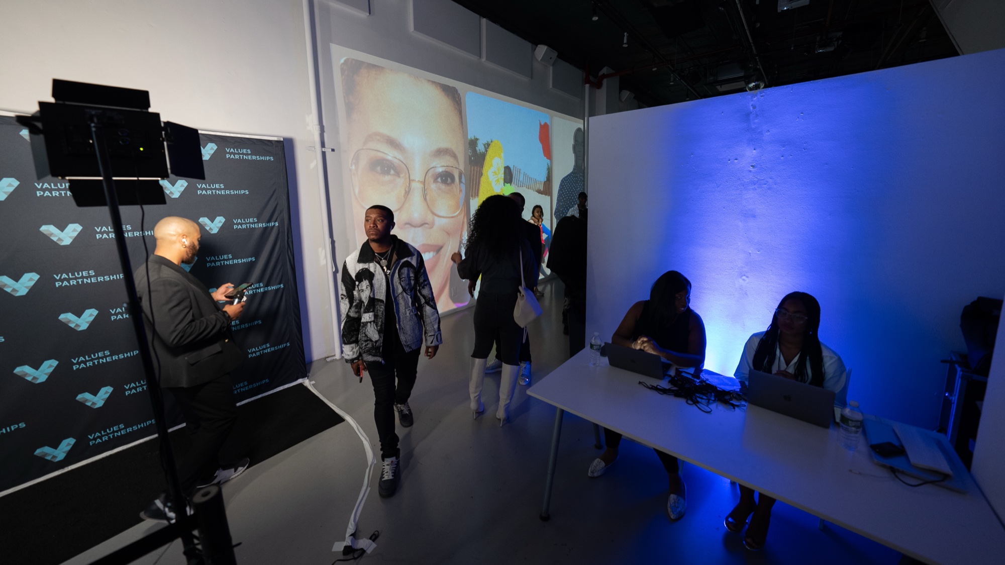 Man on his phone beside a Values Partnerships backdrop, a guest walking past a projected portrait and blue lit desk