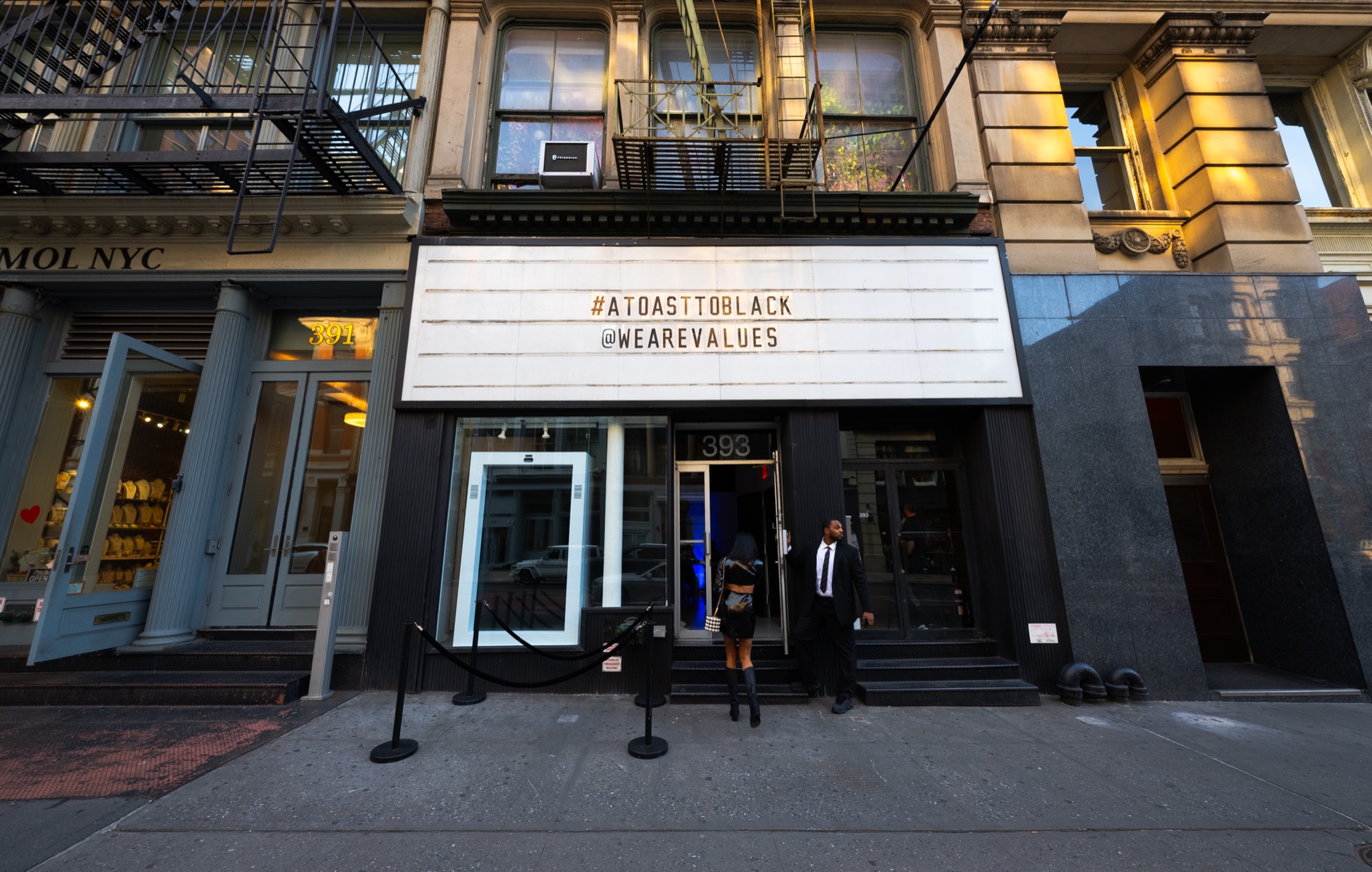 Storefront marquee showing the hashtags A Toast To Black and We Are Values above a doorman greeting a guest