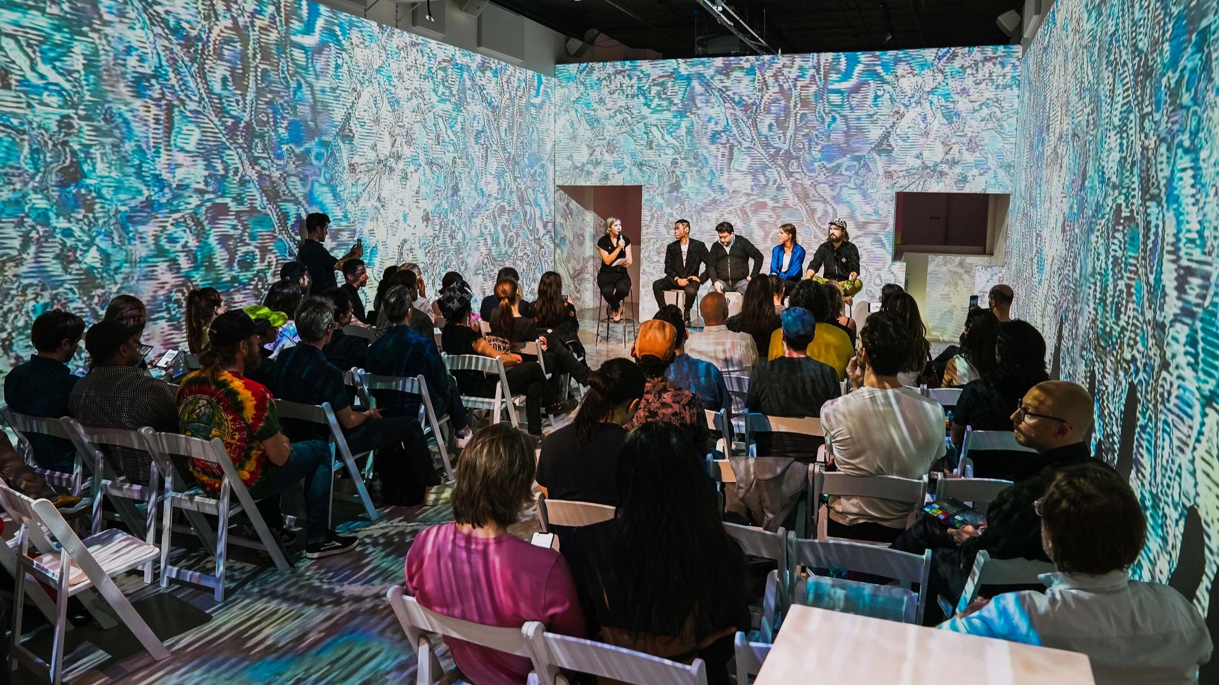Seated audience faces a panel beneath swirling monochrome projections at Vertical Crypto Art