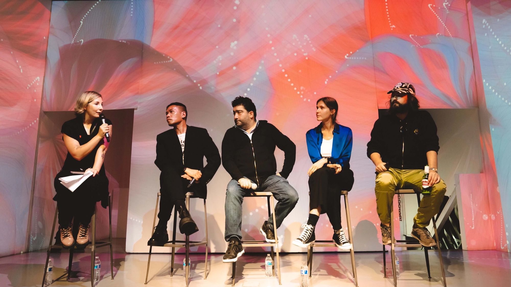 Five panelists on stools under a pink and orange projection at Vertical Crypto Art