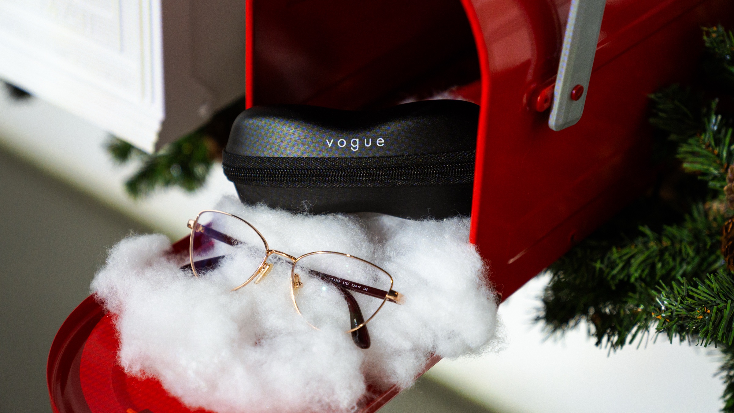 Gold rimmed glasses rest on a bed of white fluff inside a red mailbox beside a black Vogue case