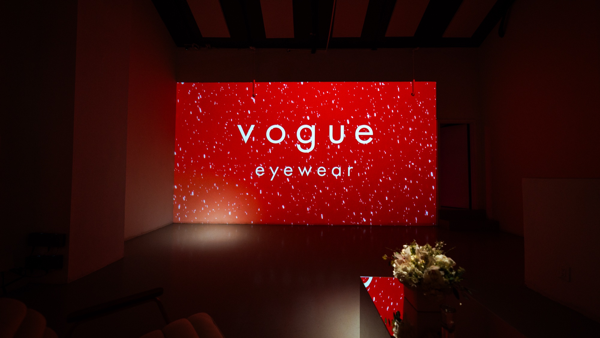 Red screen reading Vogue Eyewear speckled with white dots in a dark room, flowers on a table in front