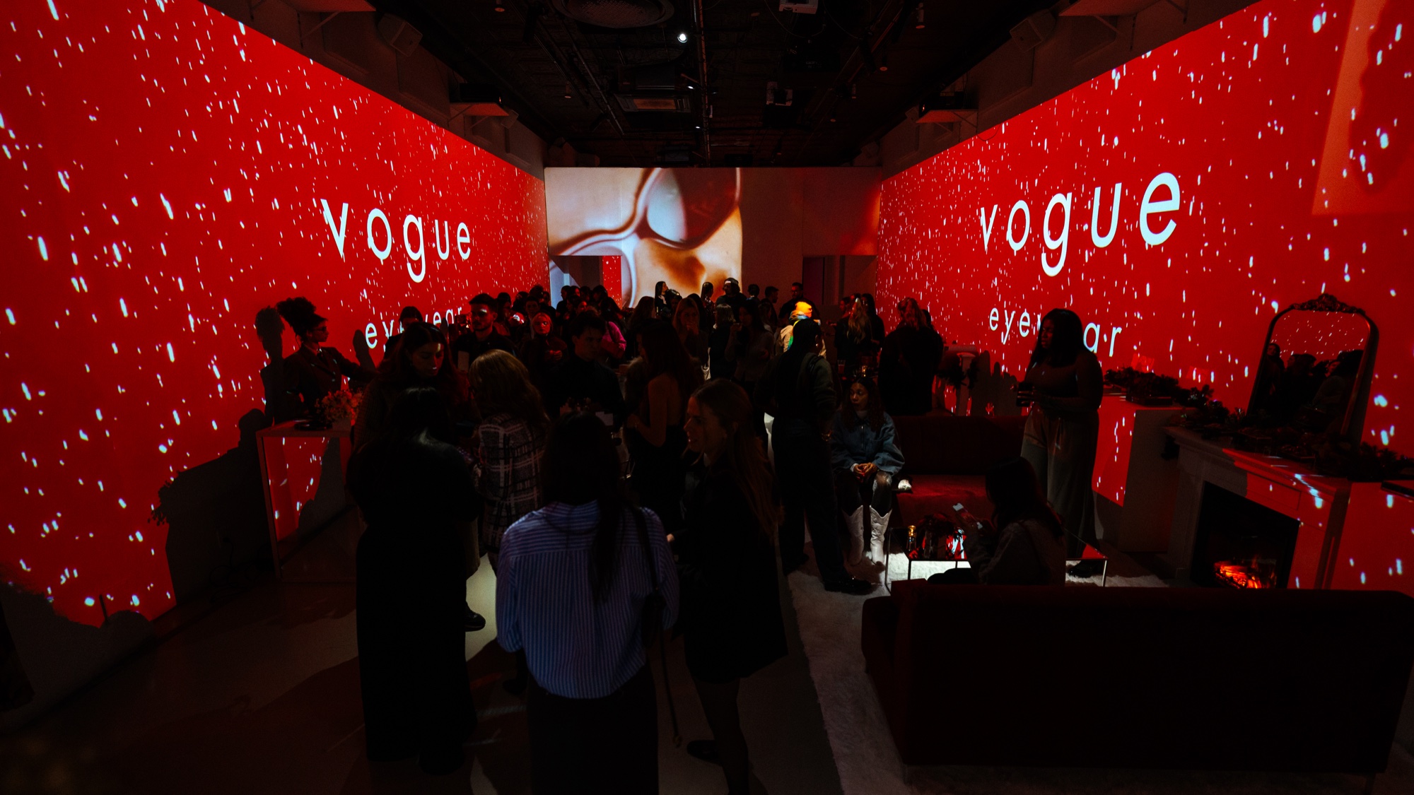 Guests mingle in a narrow room where red walls lit with white speckles carry the words Vogue Eyewear