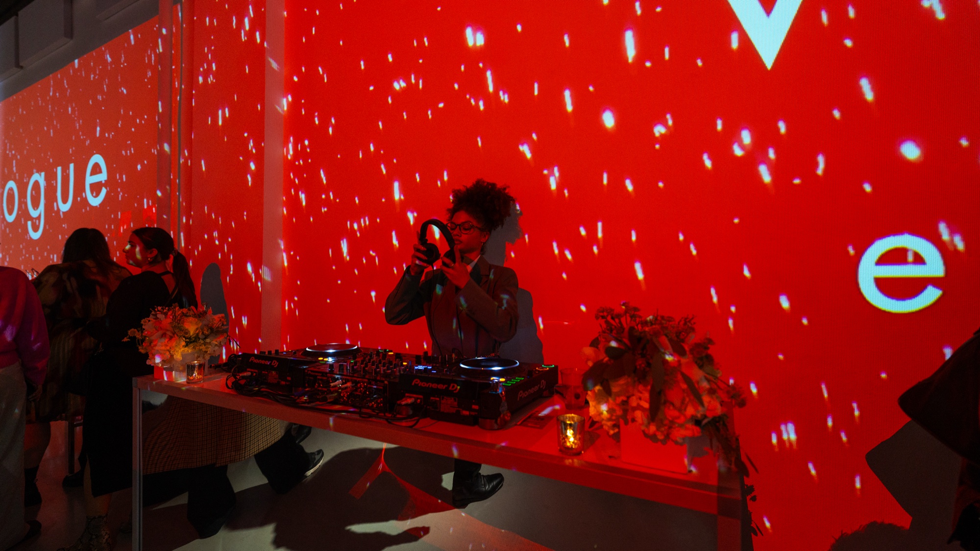A DJ with curly hair plays behind decks against a red wall projected with the word Vogue and white specks