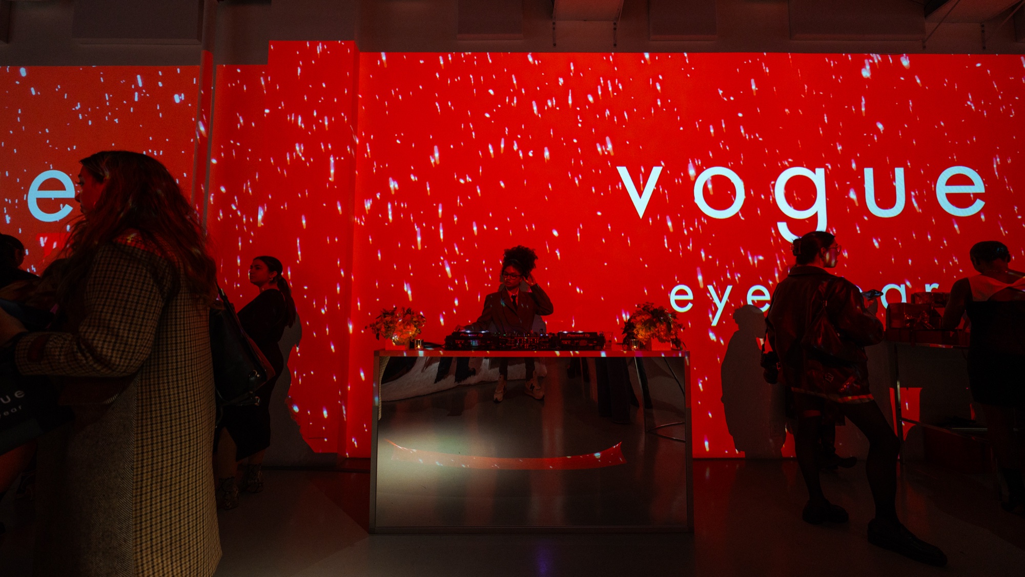 Guests seated at a mirrored counter before a red wall of drifting white specks reading vogue eyewear
