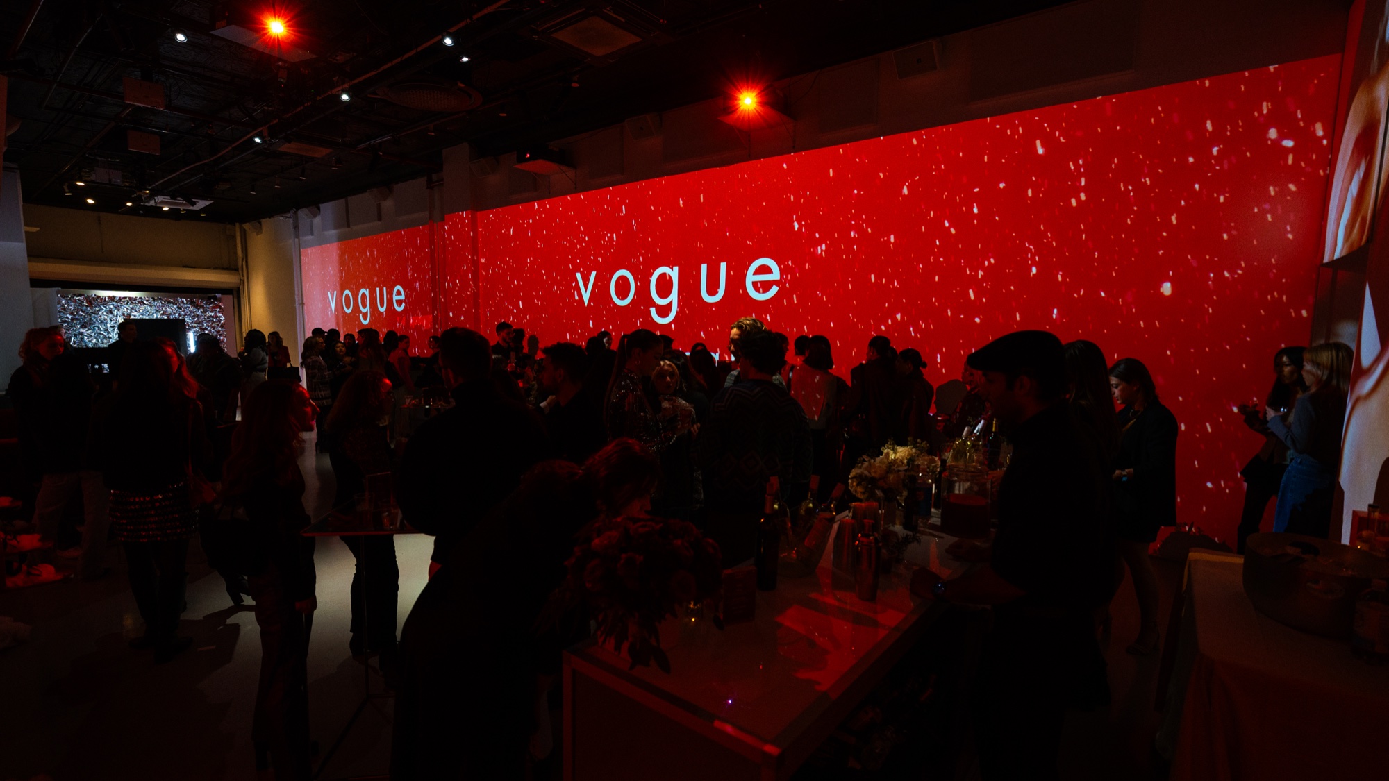 A crowd gathers under a long red wall reading Vogue, dotted with white flecks like falling snow