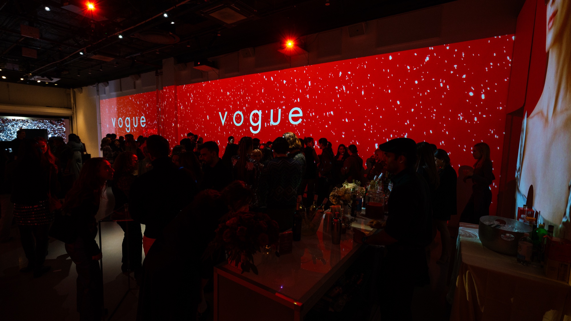 Crowd at a bar under a wide red wall reading Vogue speckled with white light