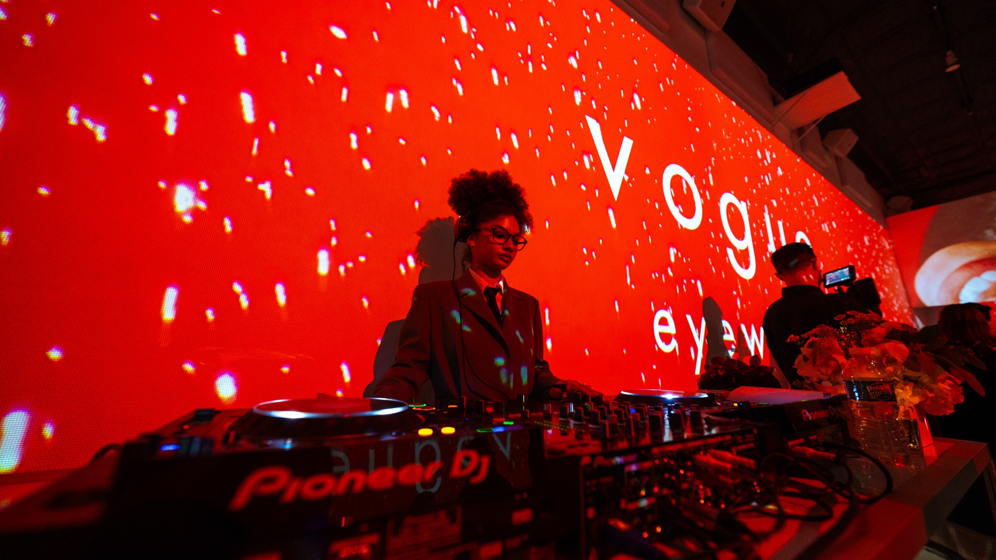A DJ in a suit and glasses works Pioneer decks before a red wall projected with the words Vogue Eyewear