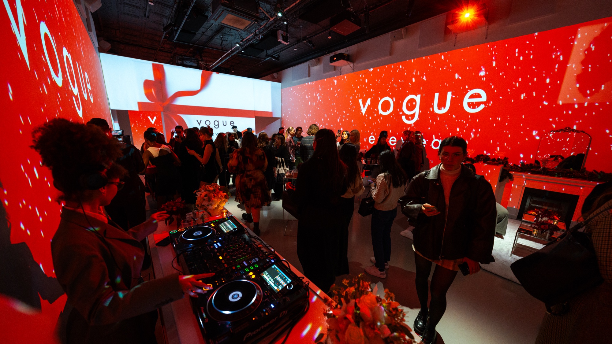 DJ decks in the foreground of a red lit room where guests mingle beneath vogue projections