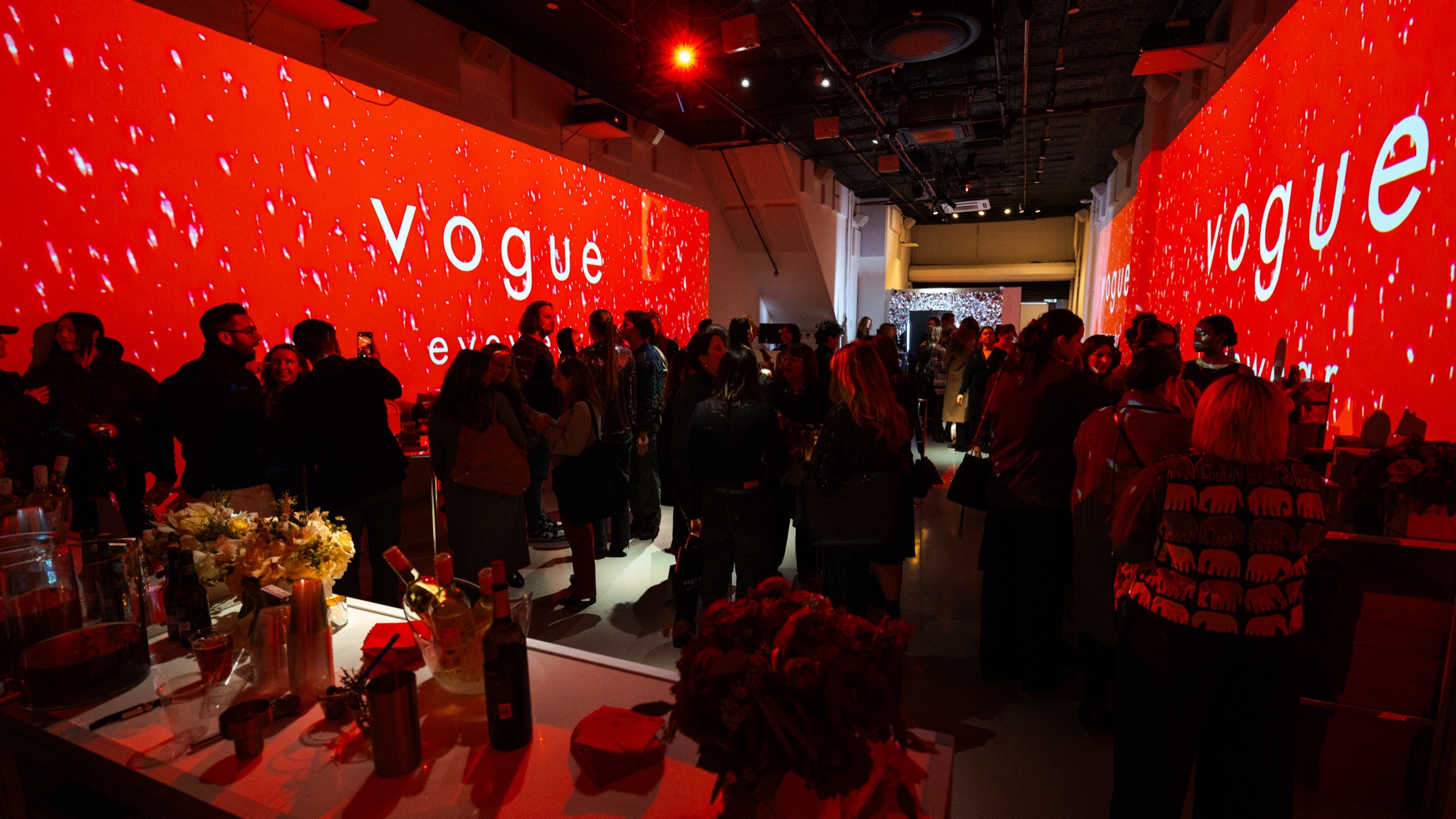 Guests chat between red projected walls reading Vogue with red roses and bottles on a table in the foreground