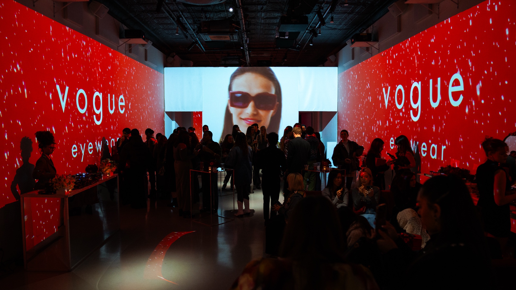 Crowd in a red lit hall facing a screen with a person in dark sunglasses, Vogue Eyewear on both walls