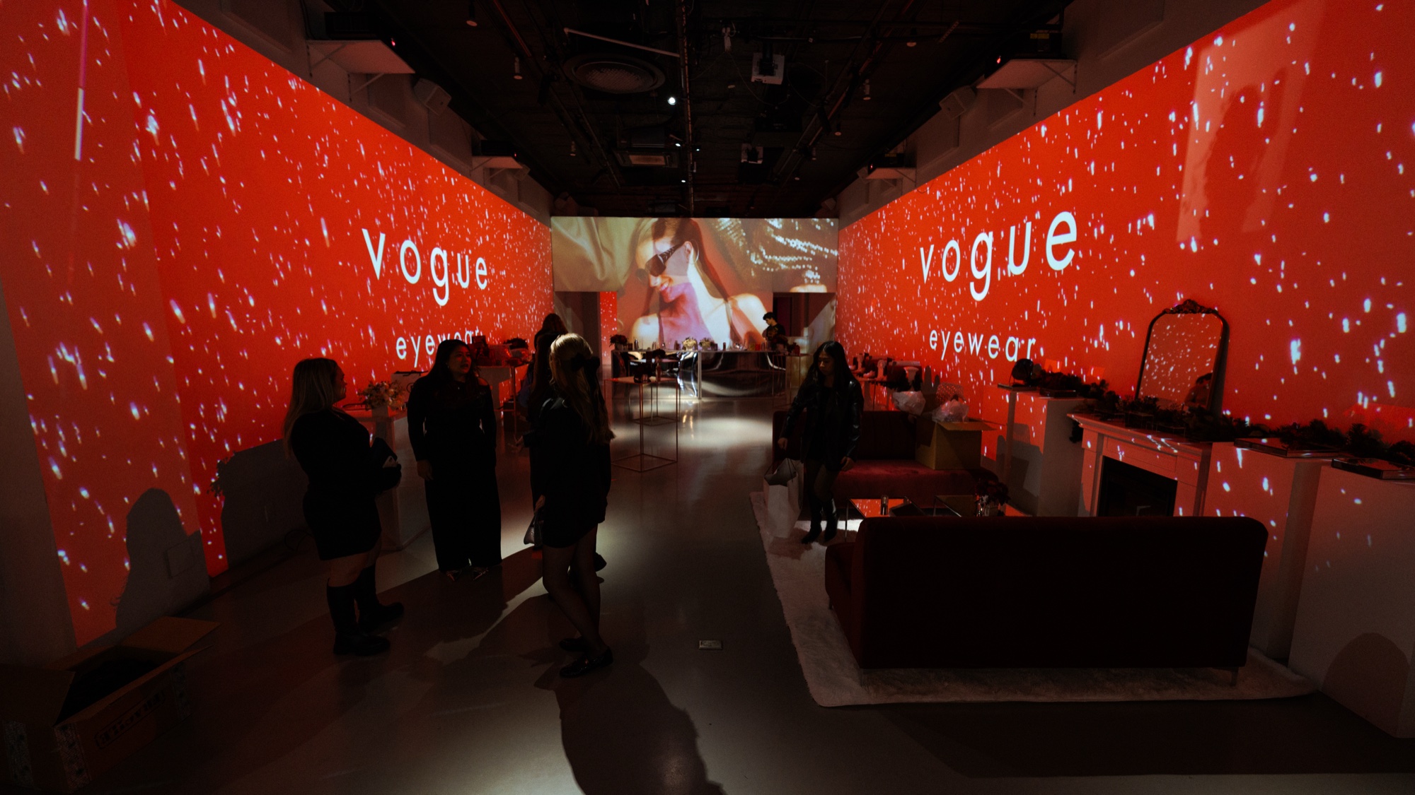 Guests in a red projected room with Vogue Eyewear on both walls and a white mantelpiece at right